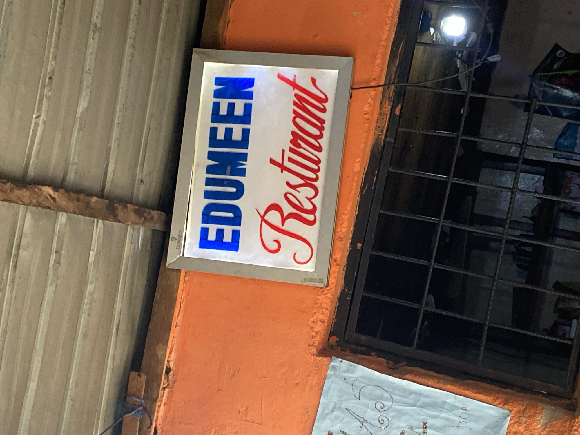 The image shows a lit sign for "EDUMEEN Restaurant". The sign is rectangular with a white background. The word "EDUMEEN" is written in large blue block letters on the left side, and "Restaurant" is written in a flowing red script on the right side. The sign is mounted on an orange-painted wall. To the left of the sign is a corrugated metal roof. To the right of the sign is a barred window. The scene appears to be outdoors, likely in an urban setting in Jos, Nigeria, given the context. There are no people visible in the image. The time of day is not explicitly stated, but the lit sign suggests it is either evening or a dimly lit area. The overall impression is of a local eatery. A small Guinness logo is visible in the bottom left corner of the sign. Below the restaurant sign and to the right, a piece of light blue paper with what appears to be decorative writing or a design is visible, partially obscured by the window bars.