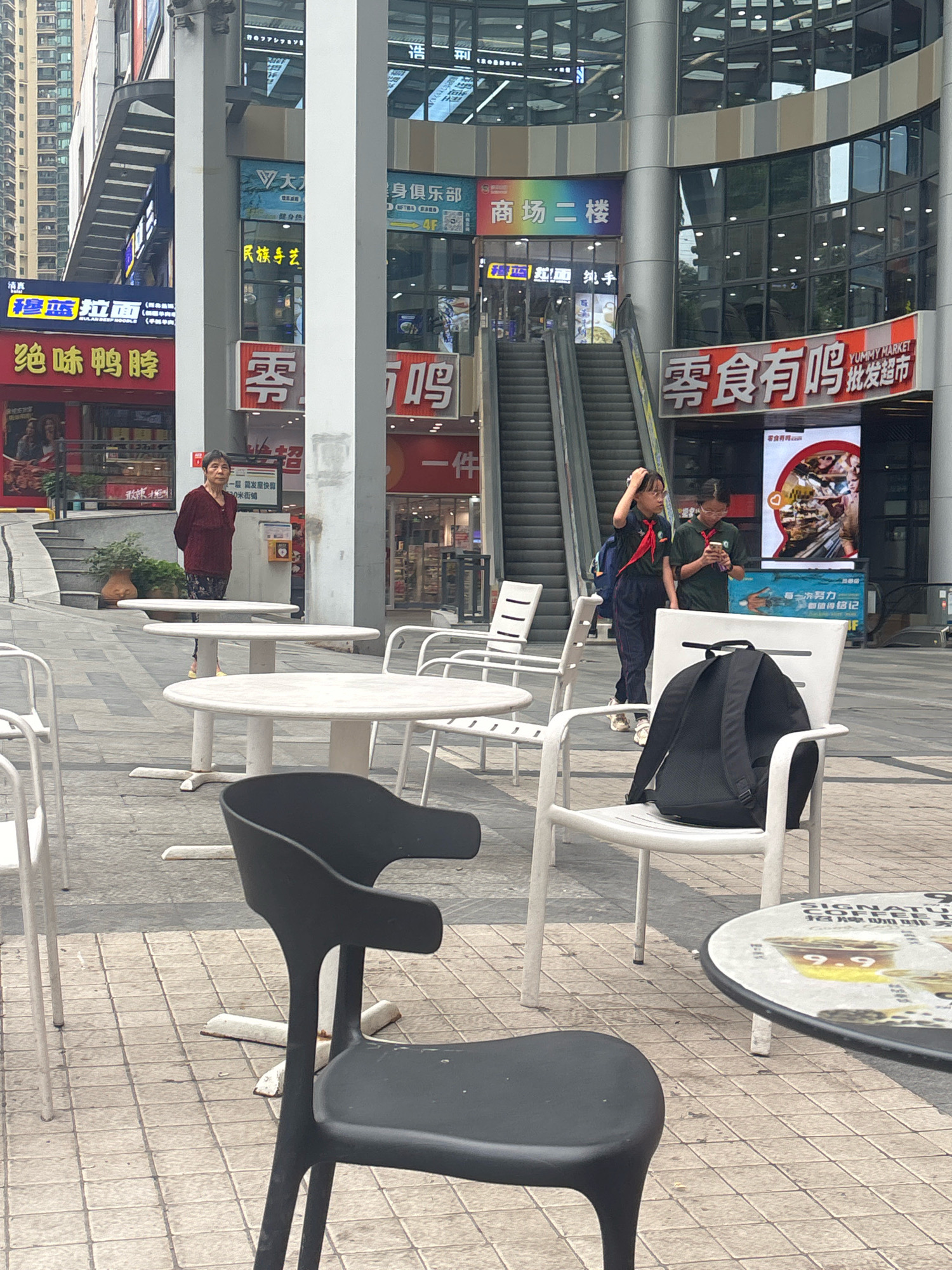 An outdoor cafe area in Nanhai District, China, features several round white tables and chairs, with one black chair prominently in the foreground. In the background, a woman stands near a table. Two young individuals, possibly students in uniforms with red scarves, are visible near an escalator. They are looking at a phone. Behind them, escalators lead to a building with multiple levels of shops and signage. The architecture is modern, with glass facades and prominent vertical pillars. One sign advertises "YUMMY MARKET WHOLESALE SUPERMARKET" and another displays food imagery. The ground is paved with square tiles. The overall scene depicts a bustling urban environment with commercial activity.
