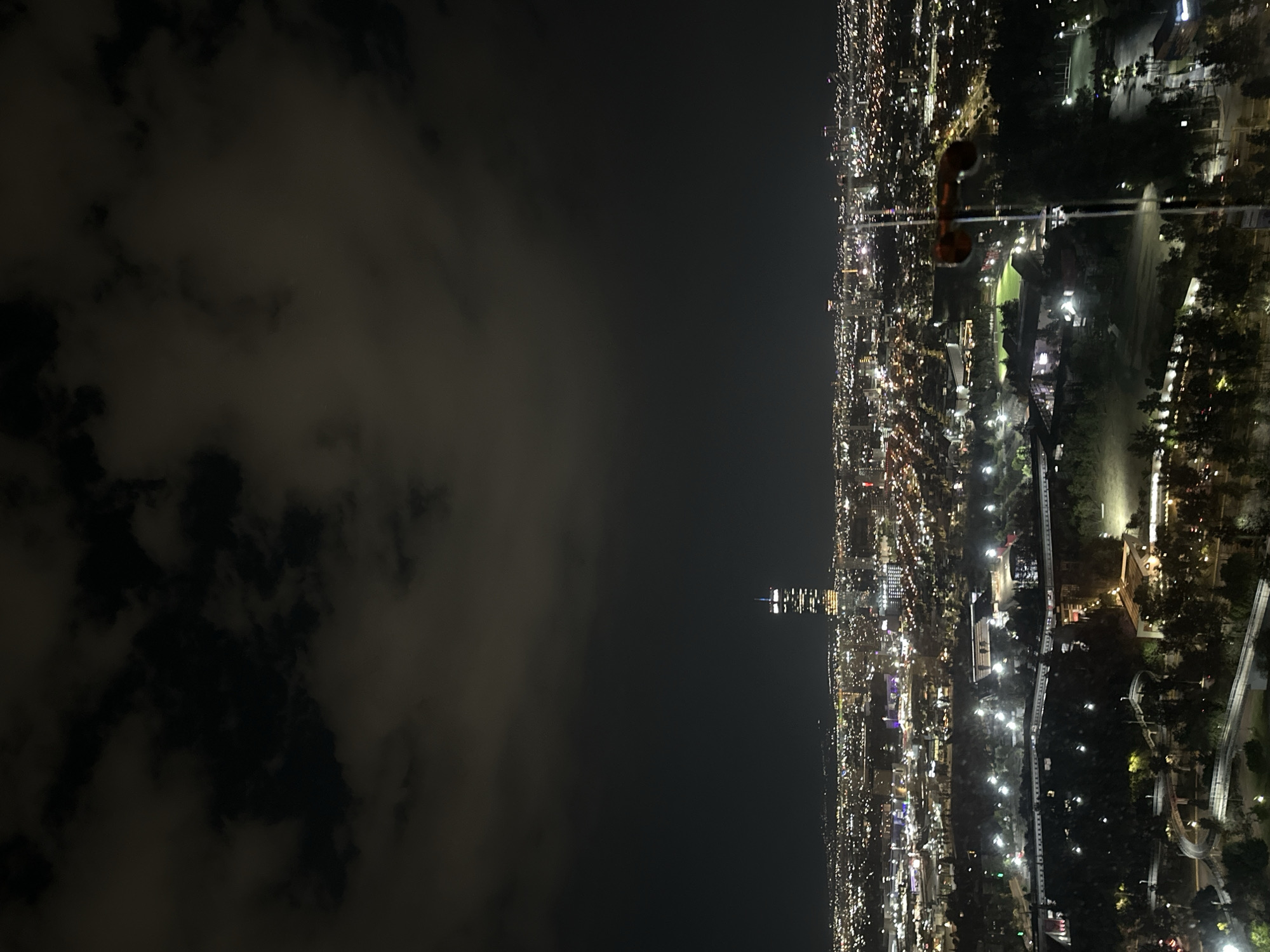 This is an elevated night view of Puebla, Mexico, specifically depicting the Angelópolis district and surrounding areas from an elevated vantage point.
The upper third of the image shows a dark night sky partially obscured by diffuse, patchy clouds. Below this, an expansive urban landscape extends to the horizon, defined by countless artificial lights emanating from buildings, streets, and infrastructure.
The prominent feature in the mid-ground is the Torre Inxignia, a tall, slender skyscraper with illuminated upper sections and interior lights, standing distinctively against the dark city backdrop. Surrounding it, the cityscape is a dense array of diffused white, yellow, and red lights from various commercial and residential structures, outlining the urban sprawl.
The lower third of the image features a well-lit park area, identifiable as part of the Parque Lineal. Key elements within this park include: a winding elevated pedestrian walkway, the *Paseo Lineal*, brightly illuminated with continuous white lights; multiple other curved pathways and ramps; several illuminated buildings with distinct architectural forms; and sections of green space, including a large, rectangular field resembling a sports ground, illuminated by floodlights. Trees are visible throughout the park, casting shadows under the artificial lighting.
In the immediate foreground, a dark, vertical mast-like structure with an attached blurred, reddish object extends upwards from the lower right corner, partially obstructing the view. No people or specific human activities are discernible.