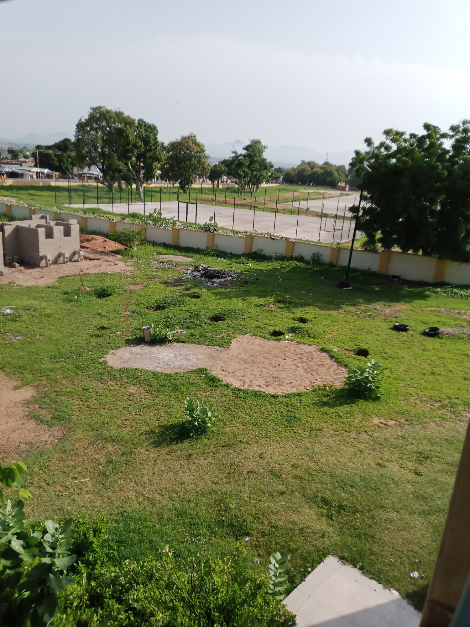 This is an aerial photograph of an outdoor area in Jalingo, Nigeria, with a focus on a partially constructed building and the surrounding grounds. In the foreground, a building made of grey cinder blocks is under construction, with several blocks laid out nearby. The ground is covered in green grass, with patches of exposed soil and a circular sandy area. There are several small bushes and some scattered debris, including two black tires and a pile of trash.
Beyond the immediate foreground, a low, yellow-topped wall with a fence runs across the middle ground, separating the grassy area from a paved court, possibly a sports field. A light pole stands near the fence. In the background, there are several trees, some with lush green foliage, and further in the distance, buildings and mountains are visible under a hazy sky. The overall impression is a developing area with both construction and natural elements.