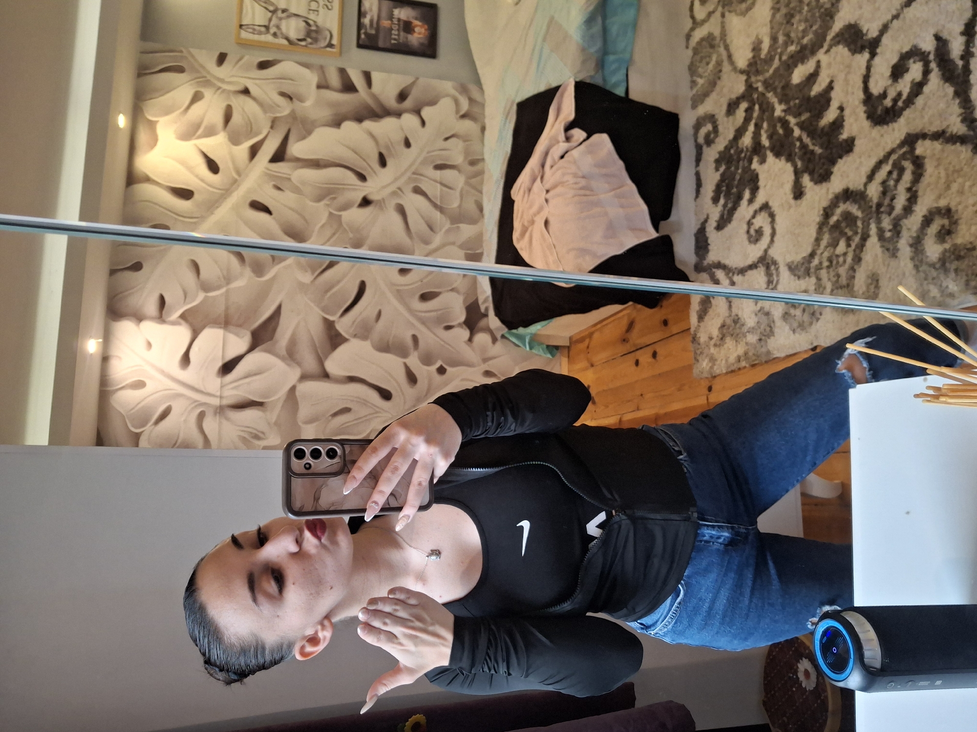 A woman is taking a selfie in a mirror. She is wearing a black sports bra with a white Nike logo, a black zip-up jacket, and blue jeans with rips. The woman has long, manicured nails with white tips. She is wearing a necklace. The background includes a wall with a large monstera leaf pattern, two framed pictures above the wall, a bed with pillows and blankets, and a rug with a floral design. There's also a wooden side table. A black soundbar is visible at the bottom right. The location is implied to be in Buhovo, Bulgaria, but there are no visual cues in the image to confirm this.