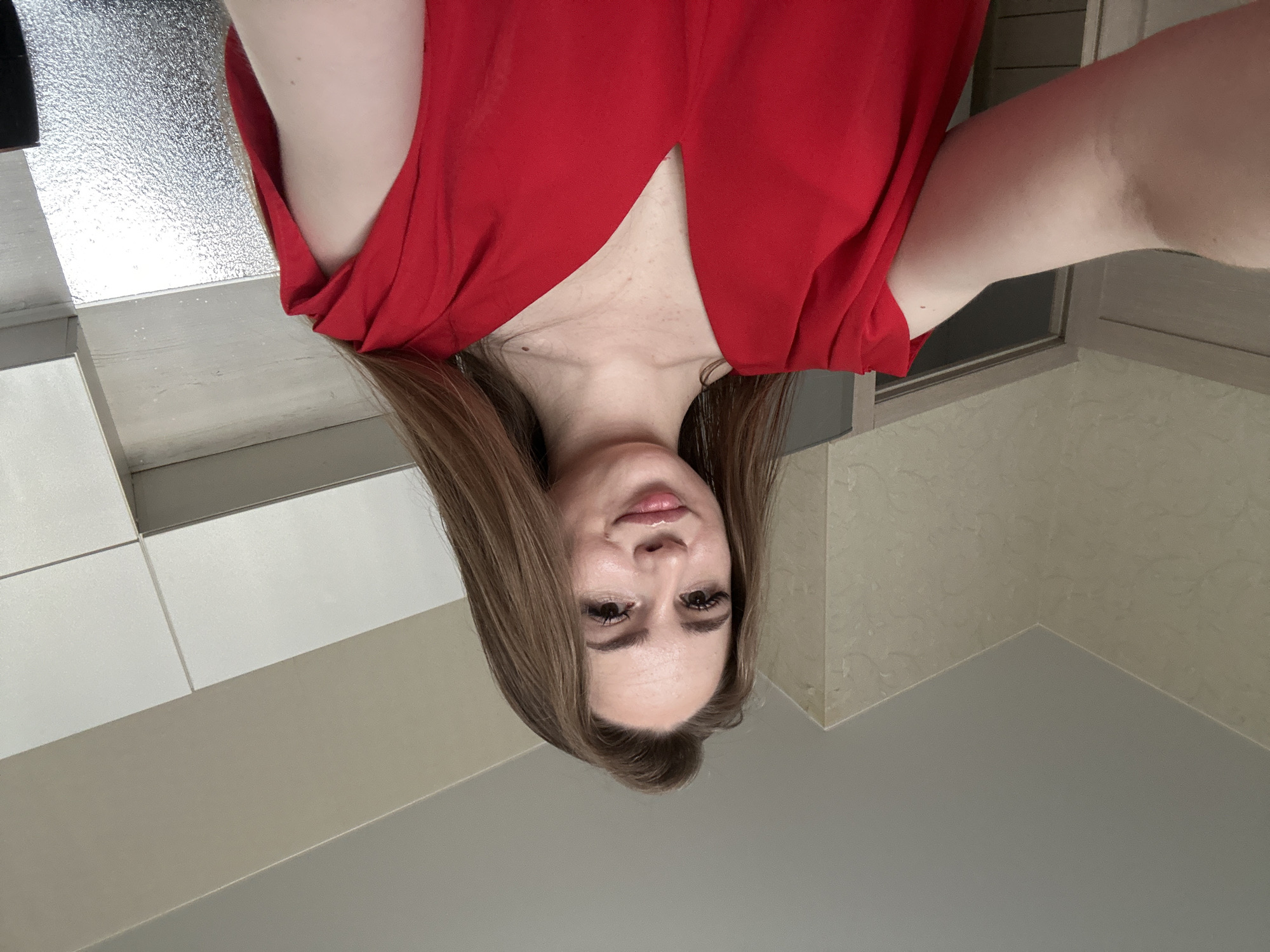 A young woman with long brown hair and fair skin takes a selfie indoors. She is wearing a bright red V-neck dress. Her left arm is extended towards the camera, and her hand is out of frame. The background appears to be a domestic interior, possibly a kitchen or hallway, with light-colored walls, cabinets, and a door visible. The lighting suggests an indoor setting with artificial light.