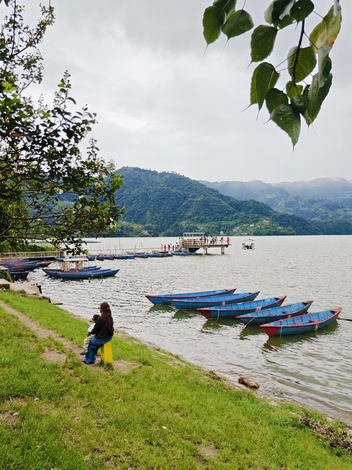 A woman and a child sit on a yellow post on a grassy bank overlooking a lake in Pokhara, Nepal. The lake is filled with numerous blue and red boats, some tied to a dock in the background where several people are gathered. Lush green hills rise in the distance under a cloudy sky. The foreground features a tree with green leaves and some white flowers.