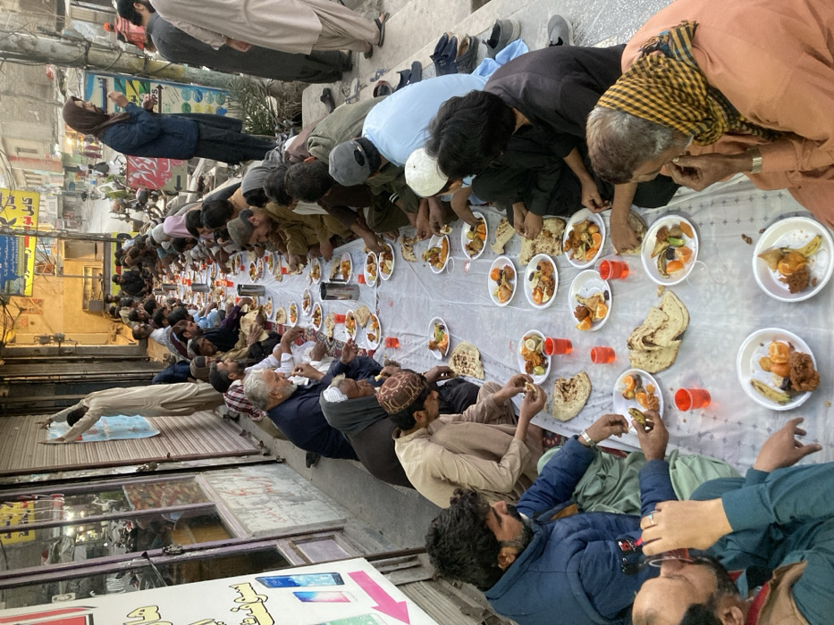 The image depicts an outdoor communal meal occurring during dusk or early evening in Quetta City Tehsil, Pakistan. Approximately 30 to 40 adult males, dressed in traditional attire including shalwar kameez and various head coverings, are seated cross-legged on the ground along both sides of a long, white plastic sheet laid directly on the pavement. Each individual has a white disposable plate containing portions of fried chicken, triangular pastries (possibly samosas), sliced oranges, and other small food items. Flatbread is placed directly on the sheet. Orange plastic cups containing a red beverage are distributed, and stainless steel thermoses or pitchers are interspersed along the length of the dining area. Individuals are actively eating, some using their hands, and drinking from the cups. Footwear is observed placed to the side of the seating arrangement. The scene is set against a backdrop of commercial buildings with roll-down metal shutters, some featuring advertisements for mobile phones. In the mid-ground, a male individual is standing, appearing to adjust or affix a blue and white poster to a closed shop shutter. The street extends into the background, showing additional structures and faint pedestrian activity.