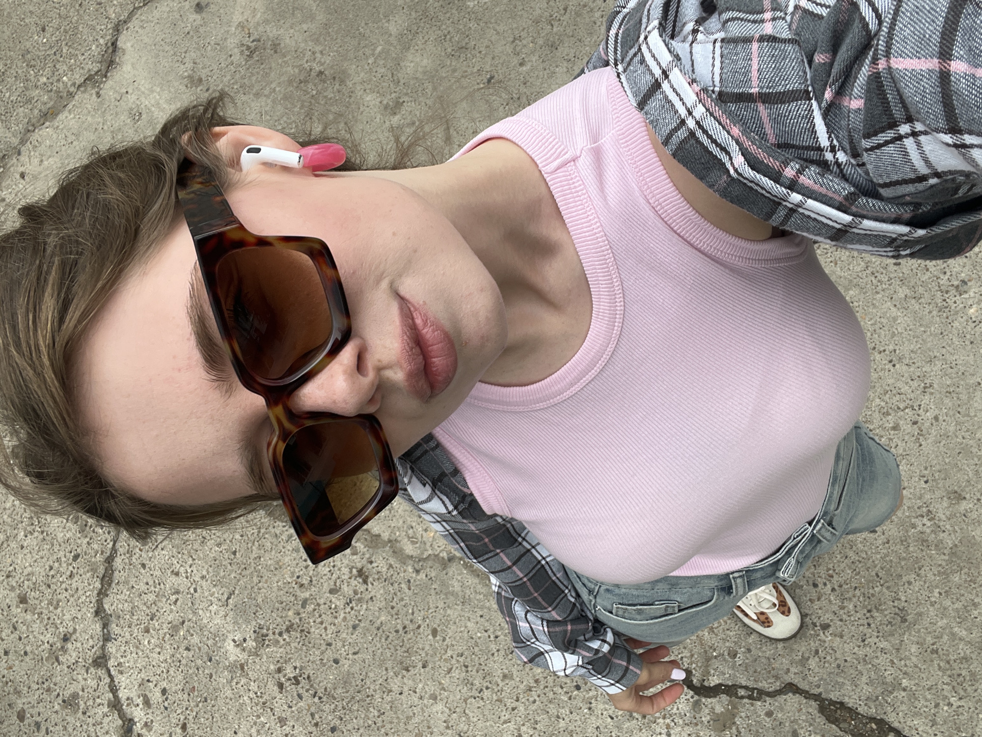 A young woman is taking a selfie outdoors on a concrete surface, possibly a sidewalk or pavement. She is wearing large, tortoiseshell-patterned sunglasses, a light pink ribbed tank top, a grey and white plaid shirt draped over her shoulders, and light-wash denim jeans. She has white wireless earbuds in her ears, with one earbud featuring a bright pink silicone cover. Her hair is pulled back loosely. The background consists of a rough, grey concrete surface with some cracks and variations in texture, typical of urban outdoor environments. There are no other people or prominent objects visible in the scene. The image appears to be taken from a high-angle perspective. There is no information in the image to suggest the location is Chaikovsky, Russia.
