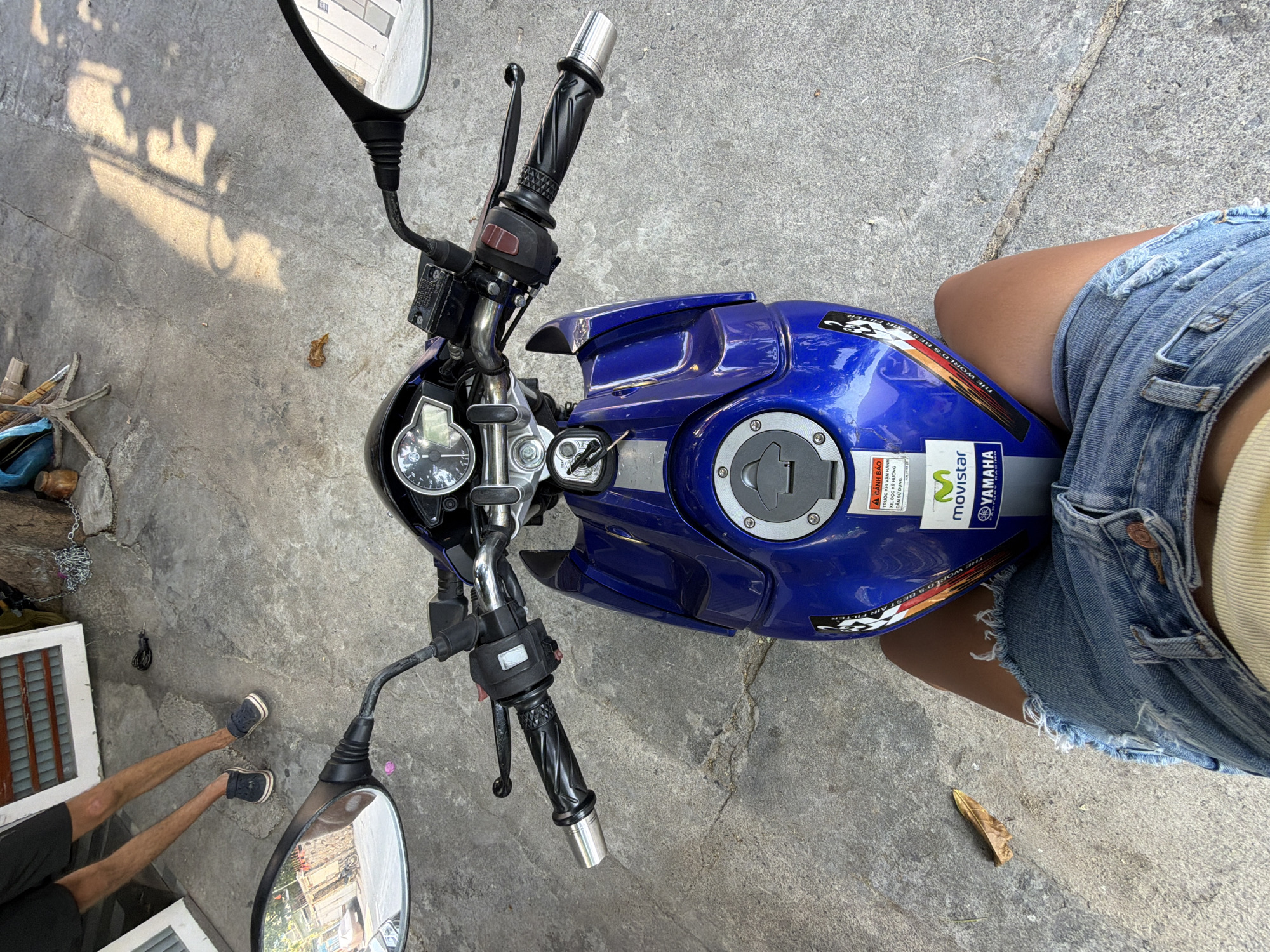 A high-angle shot shows a blue Yamaha motorcycle parked on a concrete surface. The handlebars, mirrors, fuel tank with a "Movistar" and "Yamaha" sticker, and part of the motorcycle's instrument panel are visible. A person wearing denim shorts is sitting on the motorcycle. The lower legs of another person are visible on the left edge of the frame. The photograph appears to be taken outdoors. The text on the sticker is written in Vietnamese and English. The location is in Nha Trang Ward, Vietnam.