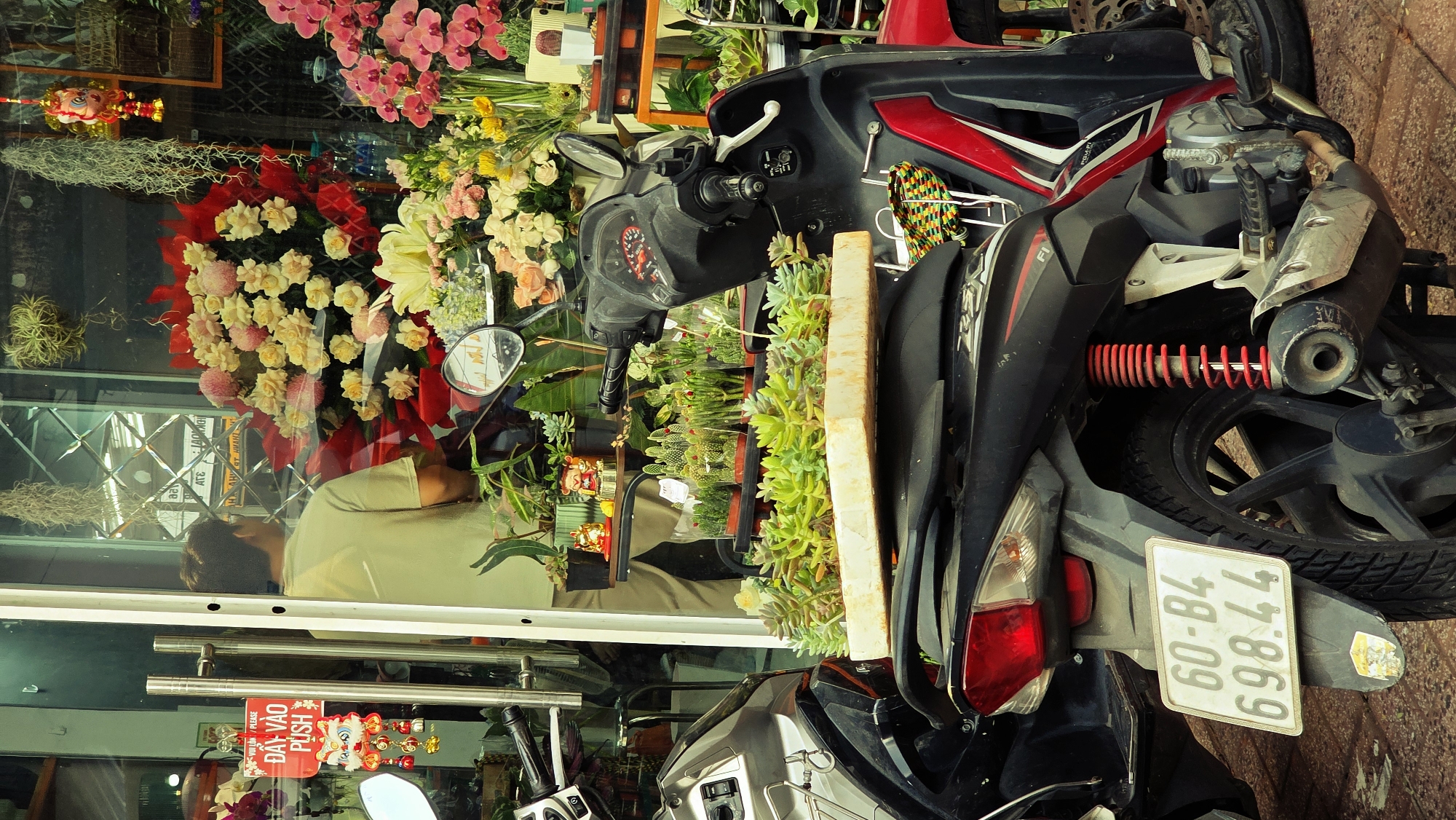 The image captures a scene in Nha Trang Ward, Vietnam, showcasing a black and red scooter parked in front of a flower shop. The scooter is carrying a wooden plank laden with small potted succulents. The scooter's license plate is visible, reading "77-B4 698.44". The flower shop's window display is filled with an array of vibrant flowers and greenery. Inside the shop, a person is seen reflected in the window, seemingly tending to the floral arrangements. The scene suggests a typical urban street setting with a business operating in close proximity to vehicular traffic.