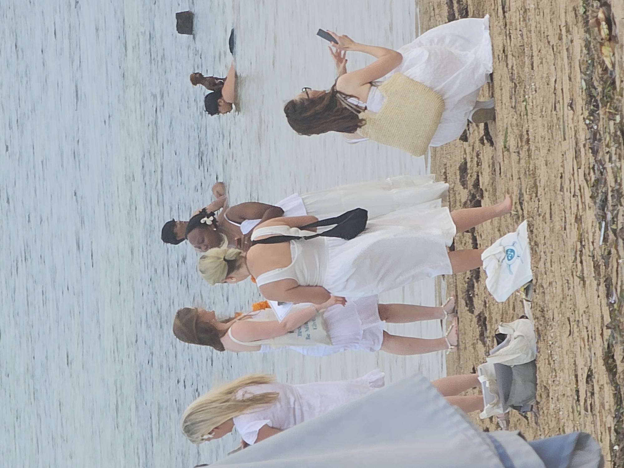 In Denpasar, Indonesia, on a beach with calm, gray water, a group of people in white attire are gathered near the shoreline. A woman in a white dress is in the foreground, holding a light brown woven bag on her lap and looking at her phone. To her left, another woman in a white dress and sandals stands with a white tote bag at her feet, while a third woman in a white dress stands further left. In the water, a man and a child are visible, along with a small brown dog perched on the man's shoulders. The sand is covered with seaweed and debris.