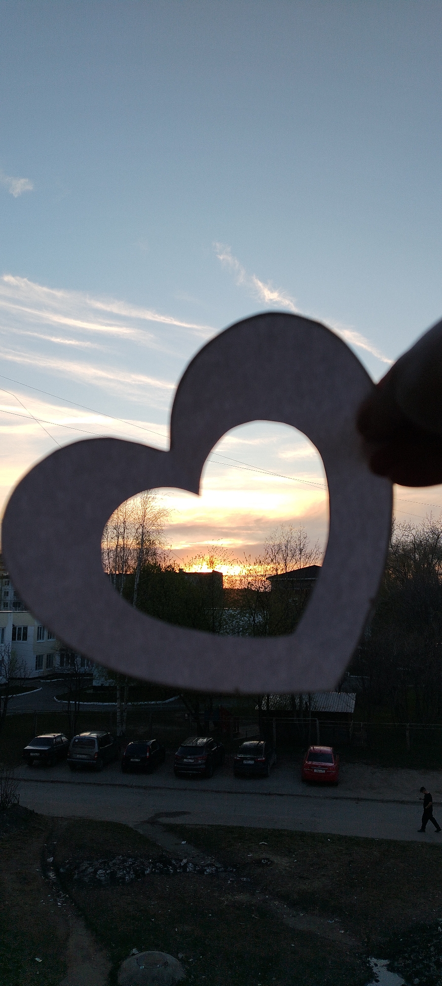 The photograph captures a sunset scene through a heart-shaped cutout. The sky is a blend of blue and orange hues, indicating either sunrise or sunset. A silhouetted treeline and buildings are visible through the heart. Below, there is a parking area with several cars of various models and colors. A pedestrian is walking on the sidewalk. The location appears to be a residential area in an urban setting.