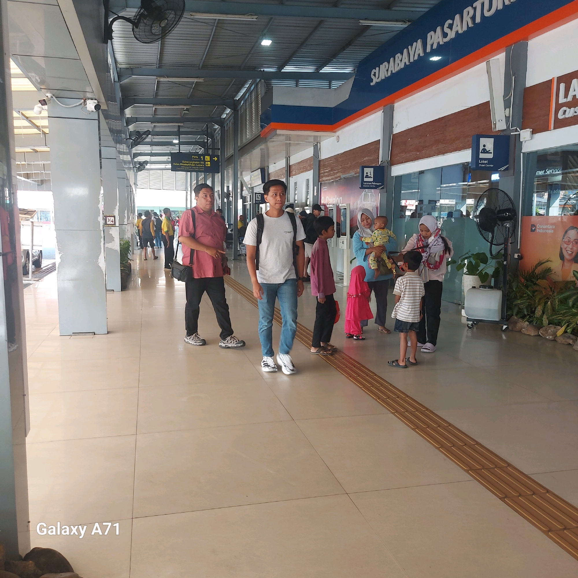The image shows the interior of the Surabaya Pasar Turi train station in Surabaya, Indonesia. It is a bright, indoor environment with a tiled floor. Several people are visible, including a man in a red shirt and black pants carrying a black bag, a younger man in a white t-shirt and blue jeans with a backpack, and a group of women and children in the background. There are signs visible, including one that says "SURABAYA PASAR TURI". A large industrial fan is also present, along with potted plants and promotional posters. The overall scene depicts a busy public transportation hub.