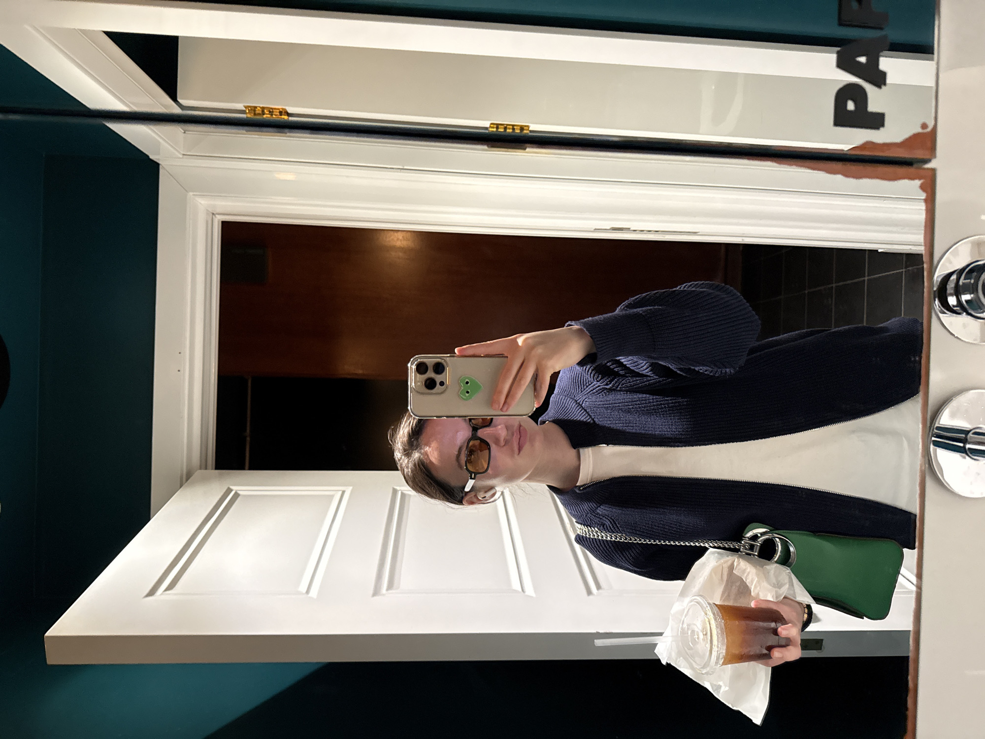 The media file, rotated 90 degrees counter-clockwise, displays a mirror selfie taken indoors. A young person, positioned upright in the frame, is looking into a smartphone held in their right hand, capturing their reflection. They are wearing a dark blue ribbed cardigan over a white top and amber-tinted rectangular glasses. Their hair is dark brown and pulled back.
In their left hand, they hold a clear plastic cup containing an iced beverage, likely coffee or tea, partially wrapped in white paper. A small, bright green shoulder bag with a silver chain strap hangs from their left shoulder. The smartphone, identifiable as an iPhone with three camera lenses, has a light-colored case adorned with a green heart sticker.
The reflection in the large mirror reveals a white, paneled door in the foreground, with a dark wooden interior or door visible beyond it. A dark teal wall is prominent on the left side of the reflection. On the right, chrome fixtures, possibly part of a door handle or mirror mounting, are visible. Partial black text, "P A R", is visible on the mirror's surface, appearing alongside a rust-colored decorative or distressed element. The setting, characterized by the ornate white door trim and gold hinges seen in reflection, suggests a well-appointed indoor space, potentially a commercial establishment in Tel-Aviv, Israel. The time of day and weather are not discernible.