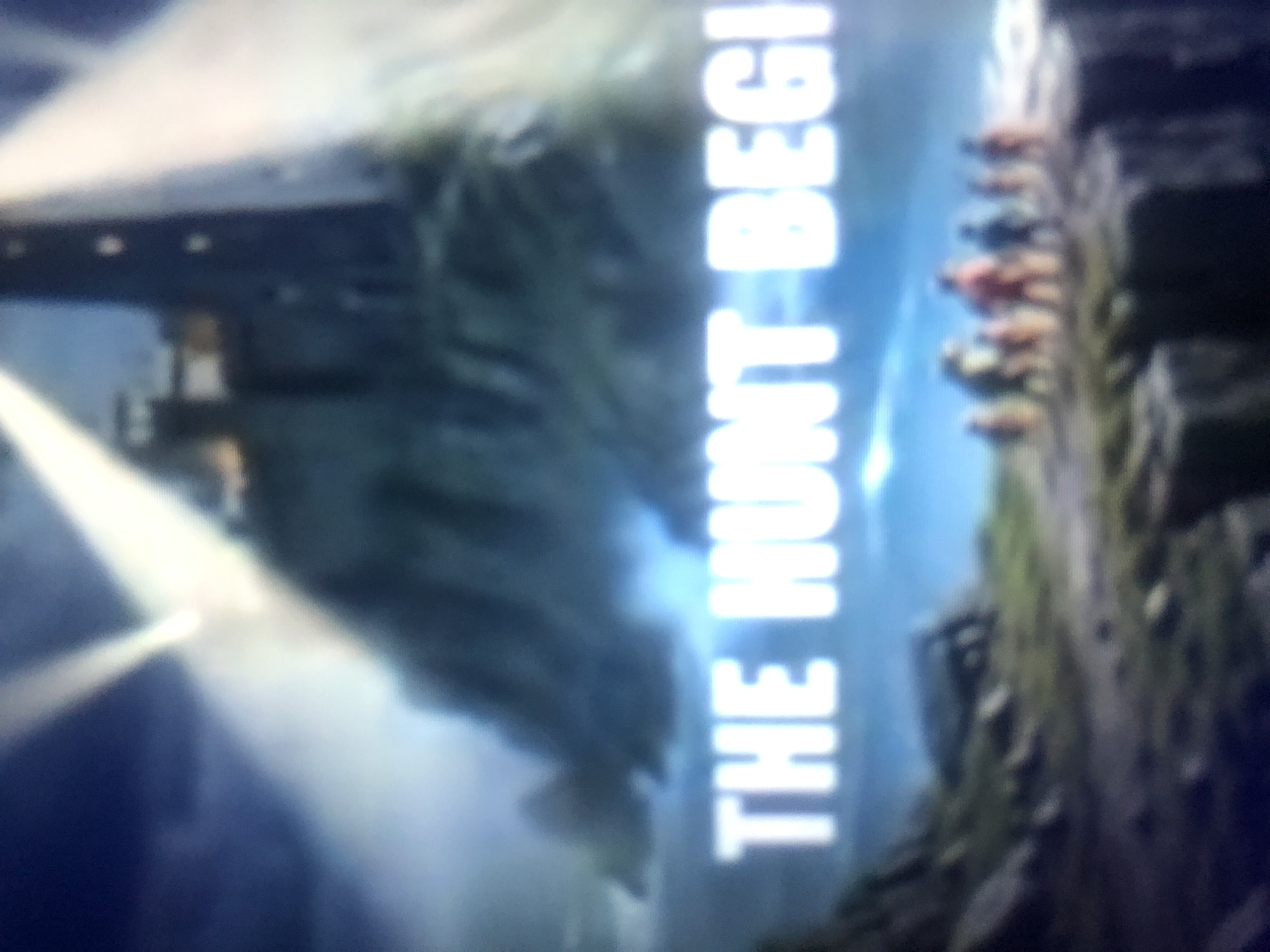 The image depicts a title card for "THE HUNT BEGI," overlaid on a dark, atmospheric scene. In the background, a tall, dark structure resembling a lighthouse stands atop a rocky, mist-shrouded island. Beams of light emanate from the top of the lighthouse, cutting through the gloom. In the foreground, a group of indistinct figures stands on the edge of a cliff overlooking a rough sea. The overall impression is one of mystery and anticipation. There is no identifiable connection to Howayi, Nigeria.