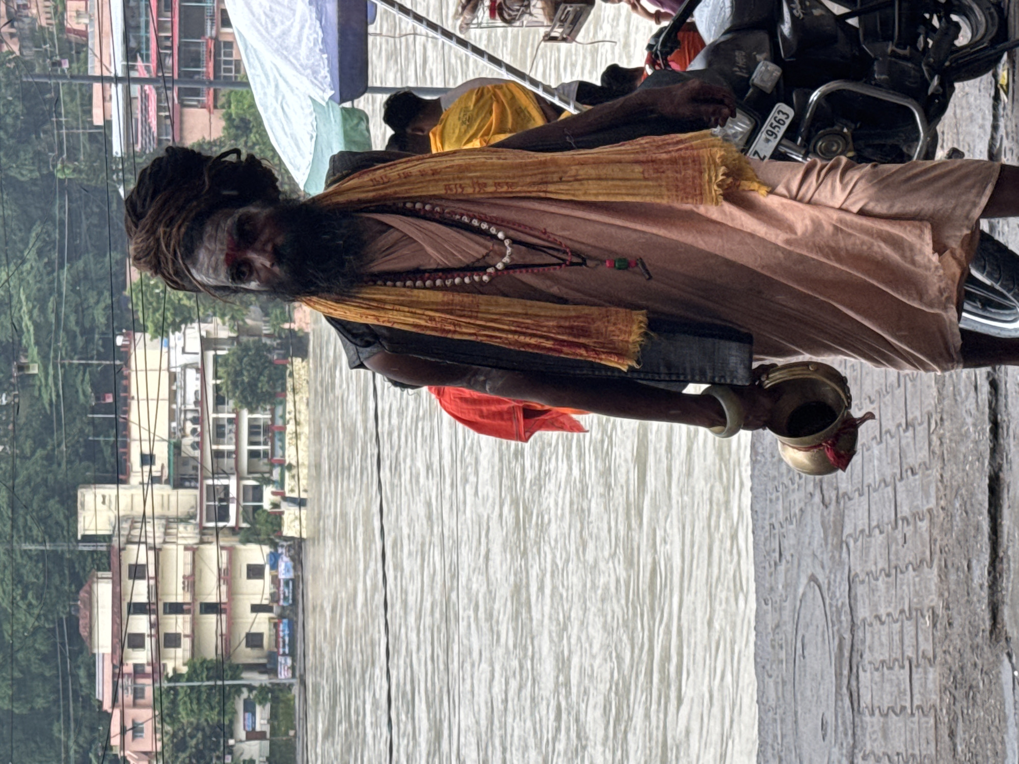 The photograph captures a scene along the Ganges River in Rishikesh, India. In the foreground, a man with dreadlocks and a beard, adorned with a red mark on his forehead and a yellow scarf, stands near the water's edge. He wears a dark vest over a peach-colored garment and holds a small brass pot. To his right, a motorcycle is parked. In the background, buildings with multiple stories line the riverbank, and the river itself is visible, with its surface rippling. Several other people are also present in the background, some near umbrellas. The overall atmosphere suggests a busy, possibly religious, location.