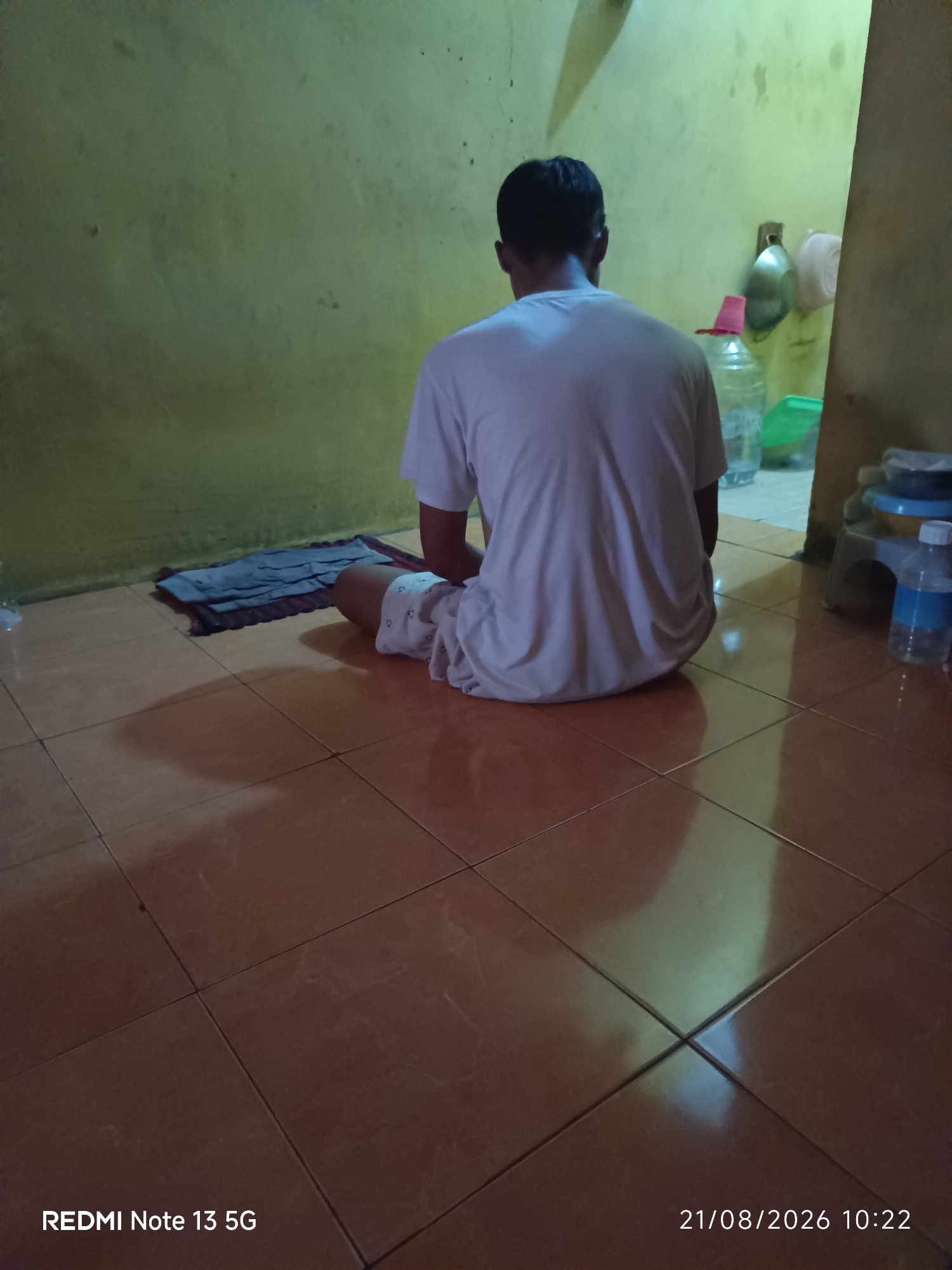 In a dimly lit room with a yellow-green wall and a tiled floor, a person is sitting with their back to the camera, facing a folded cloth on the floor. The person is wearing a white t-shirt and patterned shorts. Various objects are visible in the background, including plastic bottles, containers, and what appear to be kitchen utensils. The scene is consistent with an indoor domestic setting in Talaga Sari, Indonesia.