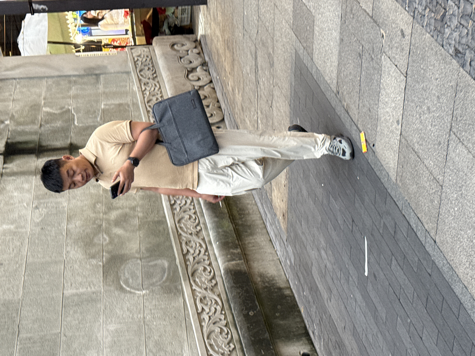 A man in a tan polo shirt and light-colored pants walks down a sidewalk in Yuzhong District, China. He is holding a smartphone in his right hand and a dark gray briefcase in his left. The sidewalk is paved with gray rectangular tiles. Behind the man, there is a stone wall with decorative carvings and a railing. In the background, a woman is visible in a brightly lit stall.