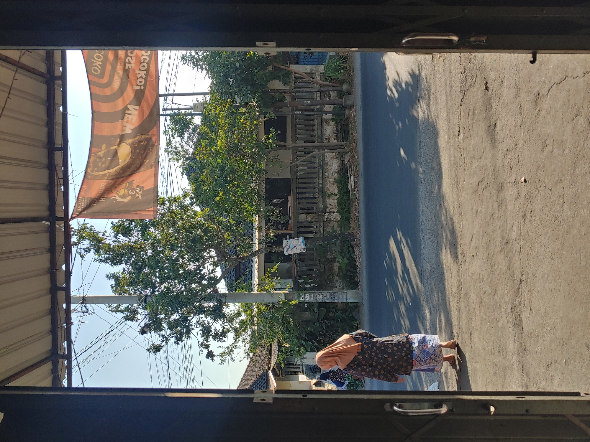 The image depicts a view through an open doorway onto a street in Kembang Kerang, Indonesia. In the foreground, a woman wearing a patterned dress and an orange headscarf walks away from the viewer on a concrete path. She is wearing sandals. The street is paved with asphalt and shows patterns of light and shadow cast by trees. Beyond the street, a low concrete fence with decorative pillars runs along the front of a residential building with a tiled roof. Trees with green foliage are situated in front of and around the house. A banner with text and images is strung across overhead. Electrical wires are visible against a clear blue sky. The overall scene suggests a warm, sunny day in a semi-urban or residential area.