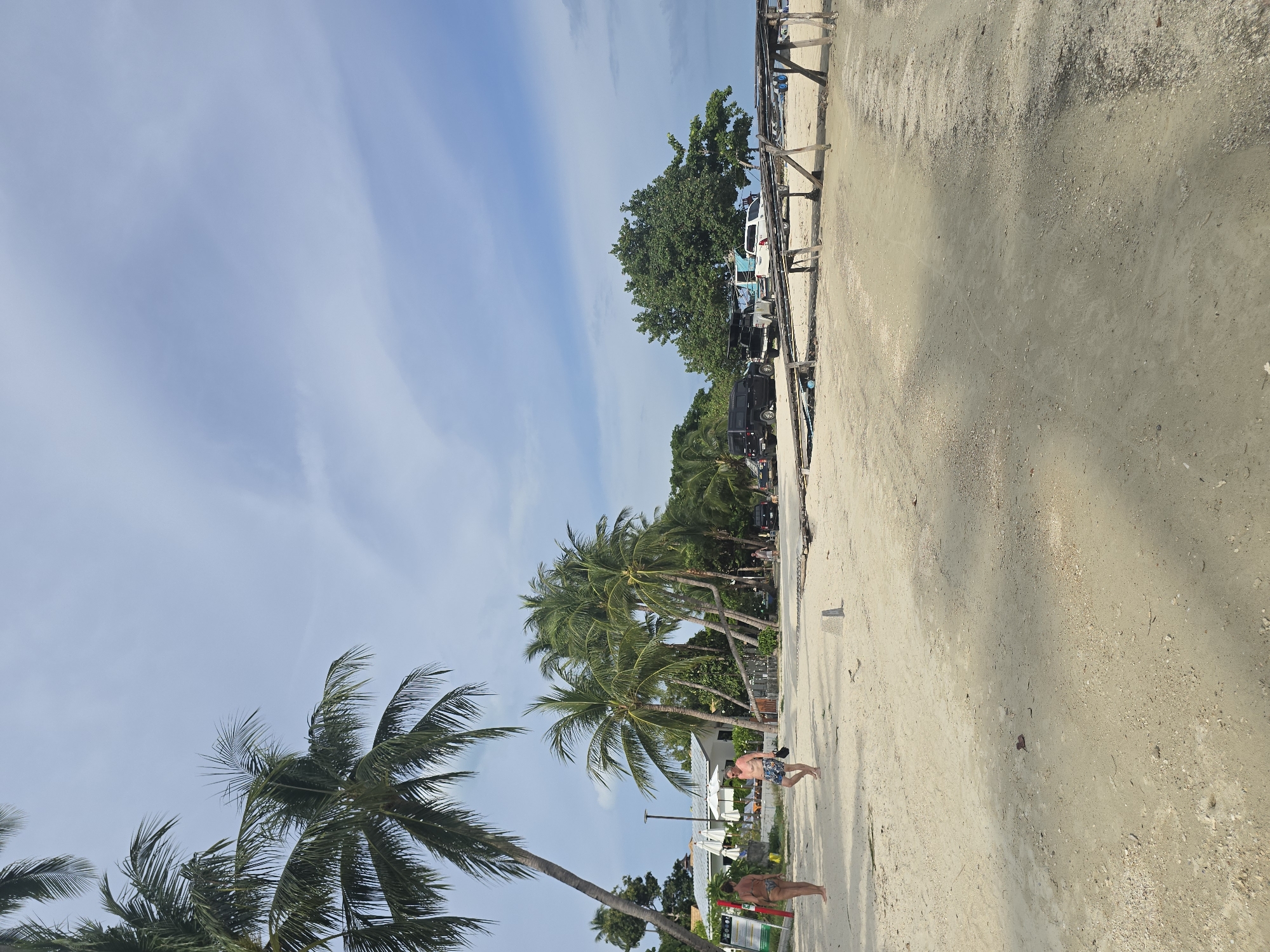 The image depicts a sandy beach with several palm trees. In the foreground, two people are walking on the sand. One is a woman in a bikini, and the other is a man wearing swim trunks and carrying a dark object. Behind them, there are more palm trees, some buildings with white umbrellas, and several vehicles, including a black SUV and a white SUV. A wooden structure is visible on the sand in the middle ground, and the sky is partly cloudy with a pale blue hue. The overall scene suggests a tropical or semi-tropical coastal environment, likely a resort area.