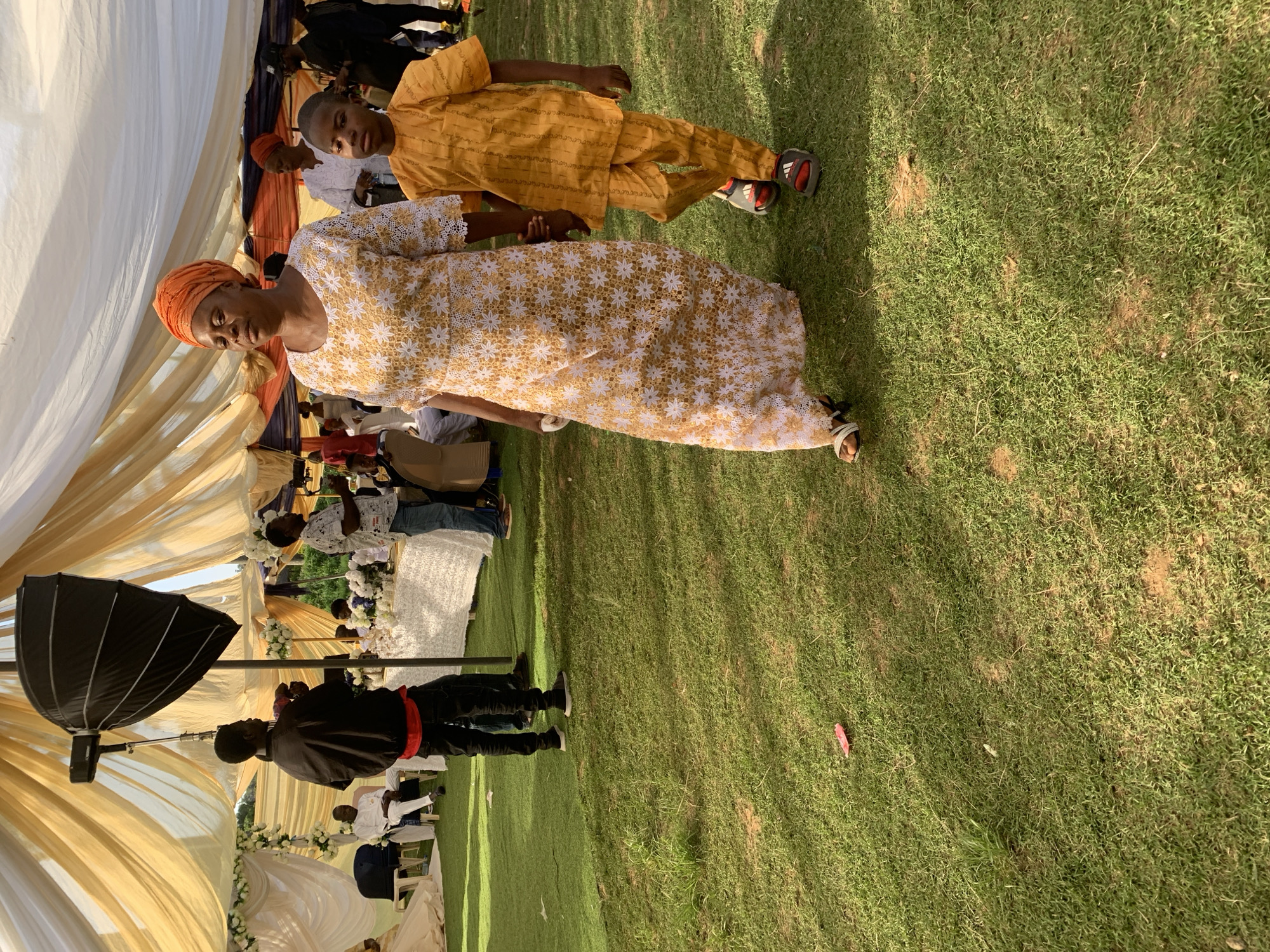 The image depicts a scene at an outdoor event, possibly a wedding, in Jos, Nigeria. A tent structure with draped fabric in cream and gold tones provides shade over a gathering of people. A woman in a white and gold lace dress and orange headwrap stands near a young boy in a yellow outfit. A photographer with lighting equipment is also visible. Other attendees are seated or standing in the background. The setting includes a grassy area.