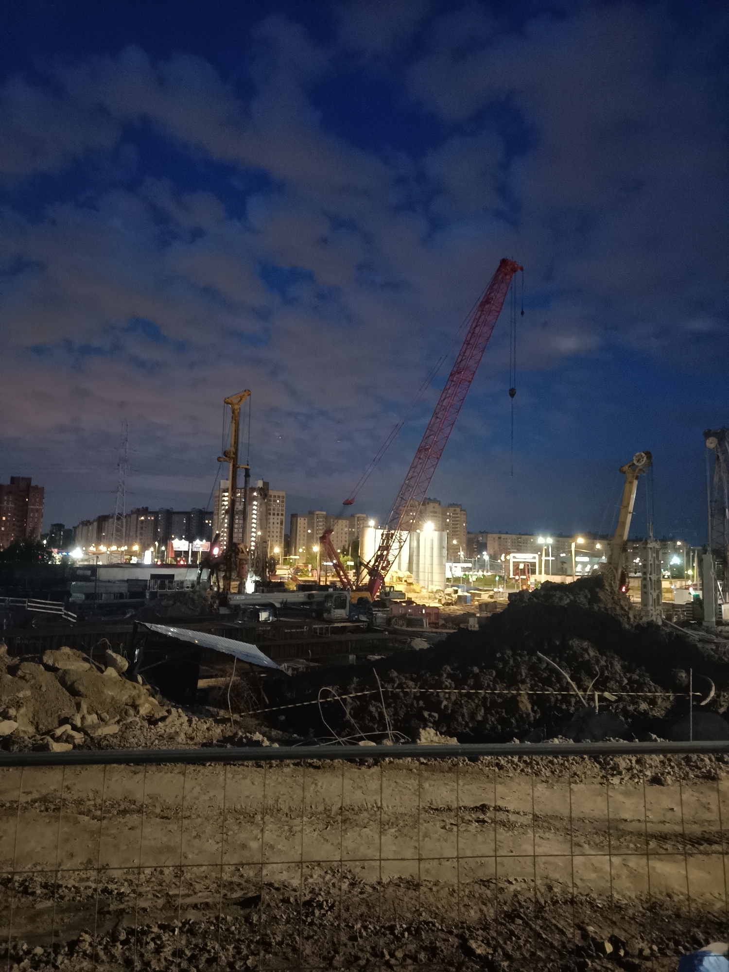 This image depicts a construction site at dusk in Saint Petersburg, Russia. The sky is a deep blue with scattered clouds, illuminated by artificial lights from the city and the construction site itself. Dominating the scene are several large construction cranes, including a prominent red lattice boom crane positioned in the mid-ground. A yellow drilling rig or pile driver is visible to the left of the red crane, along with other heavy machinery. In the background, a dense cluster of apartment buildings with illuminated windows suggests an urban residential area adjacent to the construction zone. The foreground shows excavated earth, piles of dirt, and fencing, indicating ongoing construction work. The overall impression is one of an active, large-scale construction project taking place in an urban environment during the evening hours.