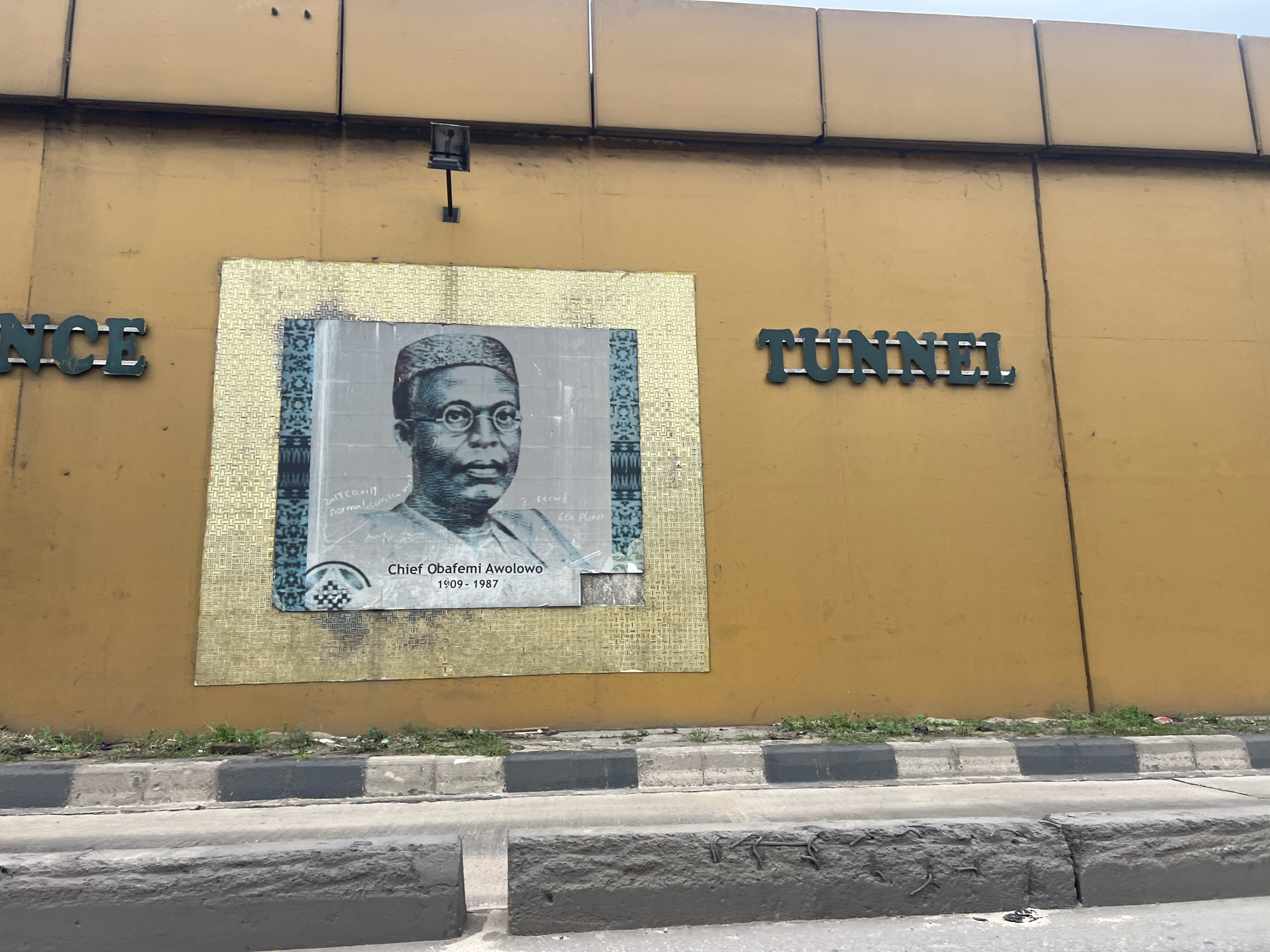 In Ikeja, Nigeria, a faded yellow wall serves as a backdrop for a large, textured mosaic featuring a portrait of Chief Obafemi Awolowo. The portrait, rendered in grayscale with blue accents, depicts Awolowo wearing a traditional cap and glasses, with his name and birth/death dates inscribed below. To the left of the mosaic, the word "ENCE" is visible in dark green, block lettering, suggesting it's part of a larger word or name. To the right, the word "TUNNEL" is displayed in similar dark green block lettering. The wall is segmented by vertical lines, indicating construction panels. A black, metallic light fixture is mounted on the wall above and to the left of the mosaic. In the foreground, a concrete curb with alternating black and gray painted sections separates the wall from a road or sidewalk area. Small patches of grass and debris are visible along the base of the wall and the curb.