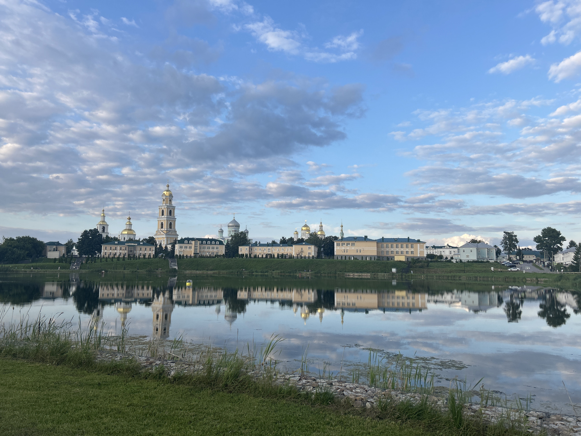 This image captures a wide-angle view of the Holy Trinity Seraphim-Diveyevo Monastery in Diveyevo, Russia, on a clear day with a partly cloudy sky. The monastery complex, characterized by its numerous golden-domed churches and bell towers, is situated on a hill overlooking a calm body of water that reflects the architecture and the sky. The foreground features a grassy embankment with some reeds and rocks along the water's edge. The buildings of the monastery are a mix of architectural styles, with prominent bell towers reaching skyward. The water's surface is smooth, creating a near-perfect mirror image of the structures on the opposite bank. Several cars are visible parked near the monastery, suggesting visitor activity. No people are clearly discernible in the scene, though the presence of cars and the manicured grounds imply human presence and maintenance. The overall atmosphere is serene and picturesque, highlighting the spiritual and architectural significance of the location.