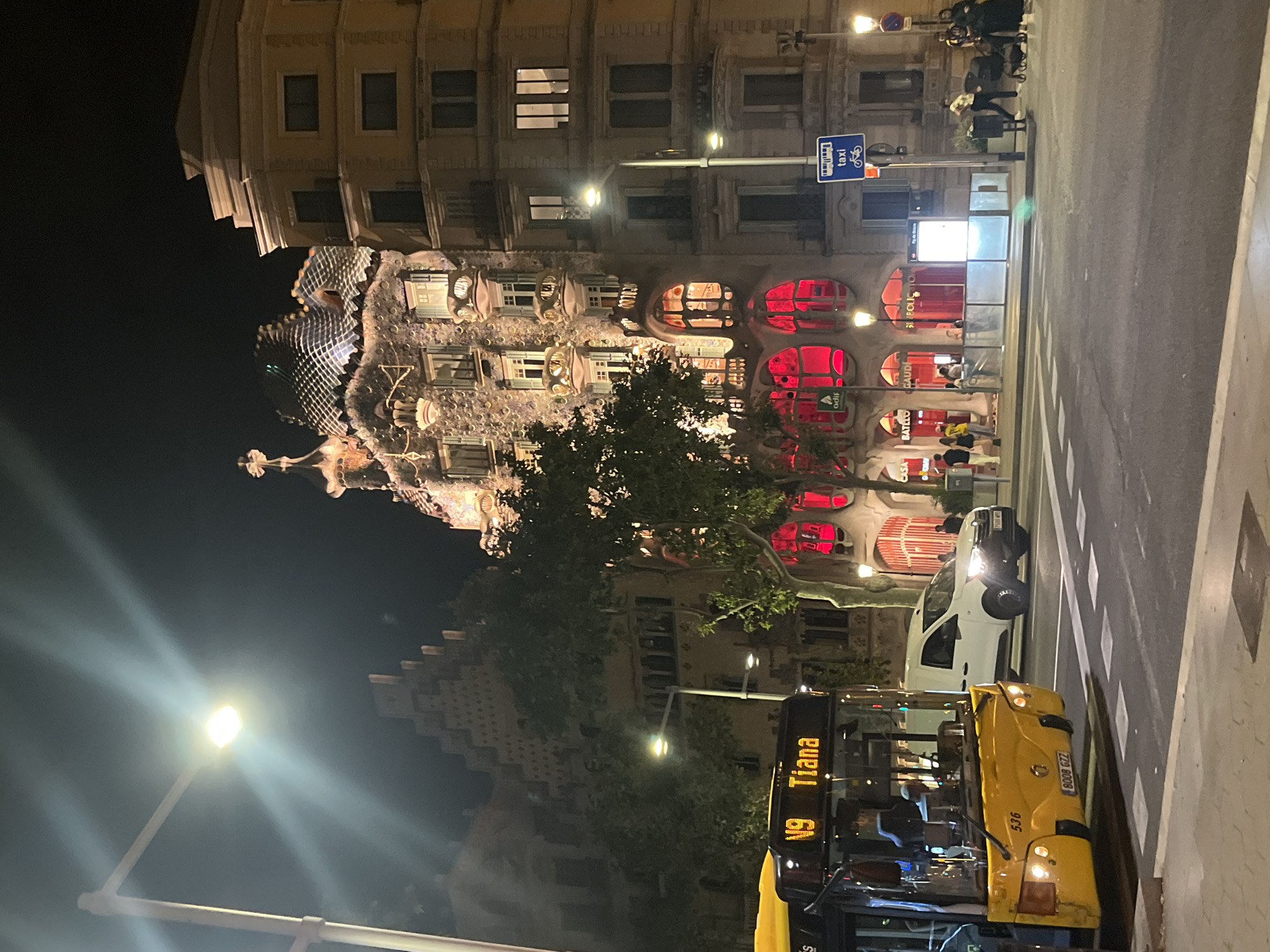 The image depicts a nighttime street scene in Barcelona, Spain, specifically in front of Casa Batlló on Passeig de Gràcia. A yellow bus, with "V9 Tiana" illuminated on its destination sign, is stopped on the left side of the frame. To its right, a white van drives by. The iconic facade of Casa Batlló, a masterpiece by Antoni Gaudí, is prominently featured in the center, its unique architectural details illuminated by the night. Red lighting emanates from the ground floor windows of Casa Batlló, creating a striking contrast with the building's textured exterior. Adjacent to Casa Batlló, on the right, is a more conventional, cream-colored building. Pedestrians are visible on the sidewalk in front of Casa Batlló and near a taxi stand sign. Streetlights are on, illuminating the urban environment. The street is paved, and a crosswalk is present in the foreground. The overall atmosphere is urban and vibrant, characteristic of a popular tourist area at night.