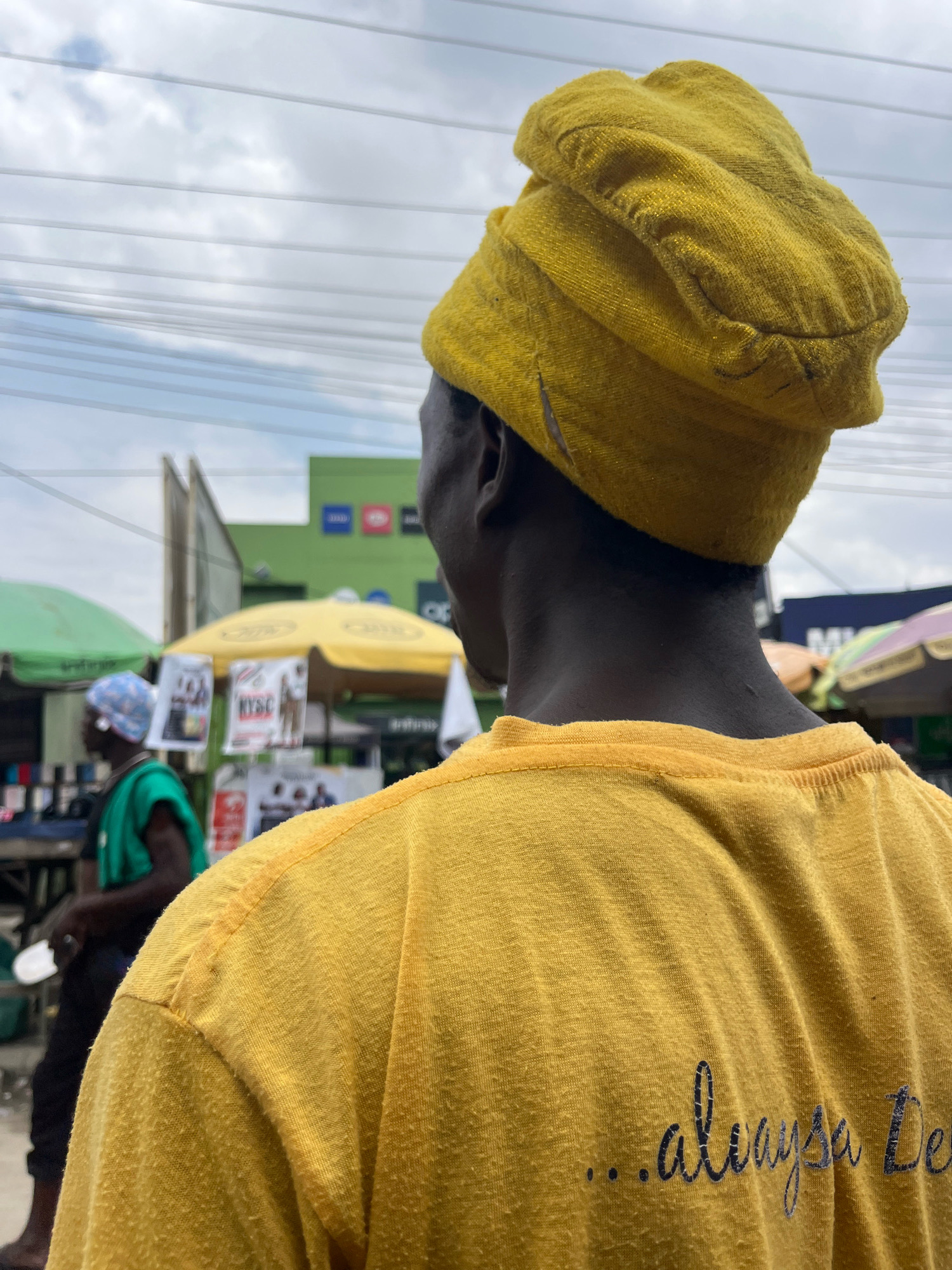 The image shows a person from behind, wearing a yellow headscarf and a yellow t-shirt with the text "...alwaysa De..." printed on it in cursive. The person is standing in what appears to be an outdoor market in Ajah, Nigeria. In the background, there are stalls with yellow and green umbrellas, advertisements, and other people. The sky is cloudy.