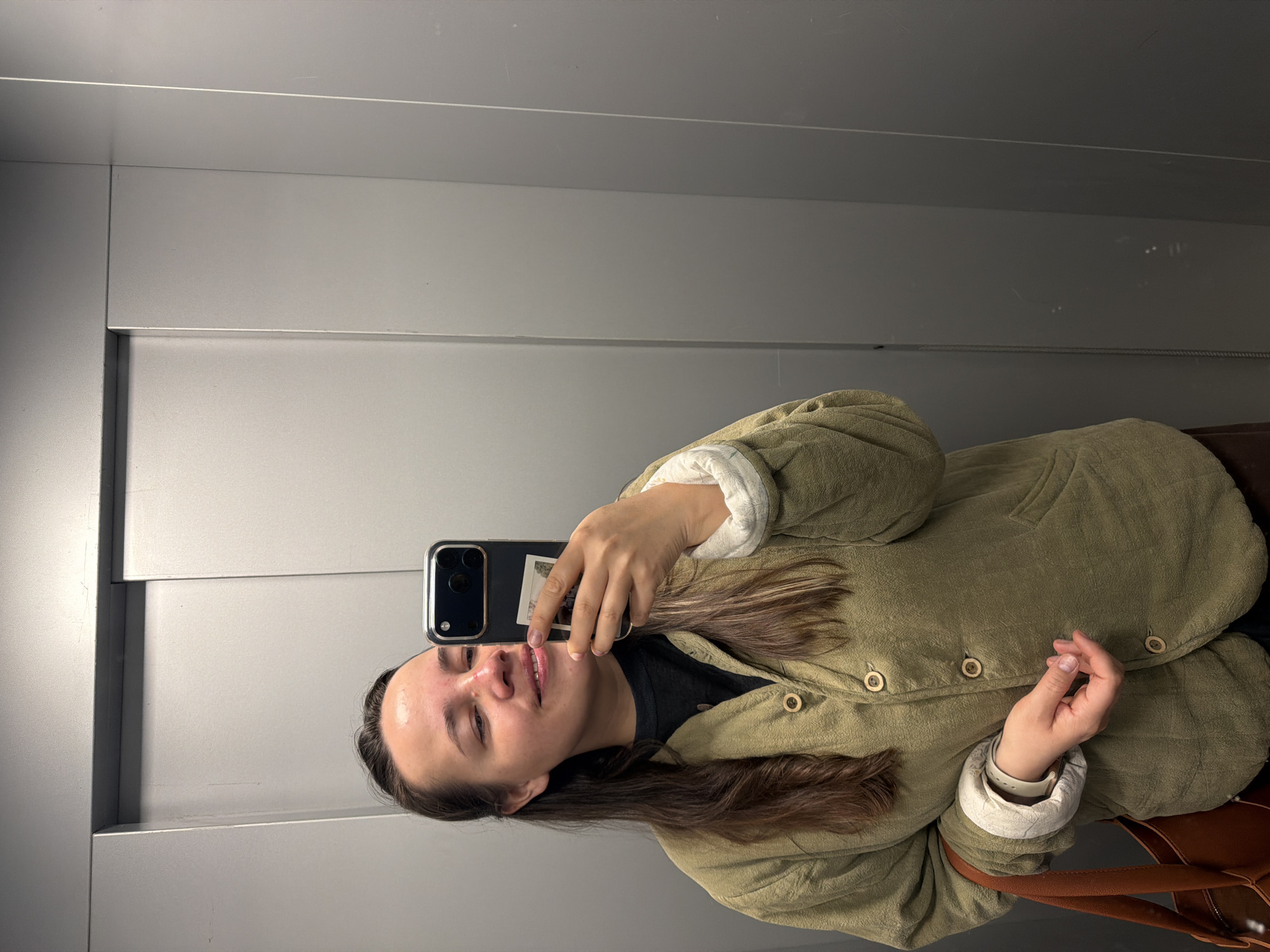 A woman is taking a selfie in what appears to be an elevator, based on the background of grey metal panels. She is holding a smartphone in her right hand, which partially obscures her face. She is wearing an olive-green jacket with button closures and white quilted lining visible at the cuffs and collar. The image is taken indoors. The city of Chaikovsky, Russia, is not evident in the image.
