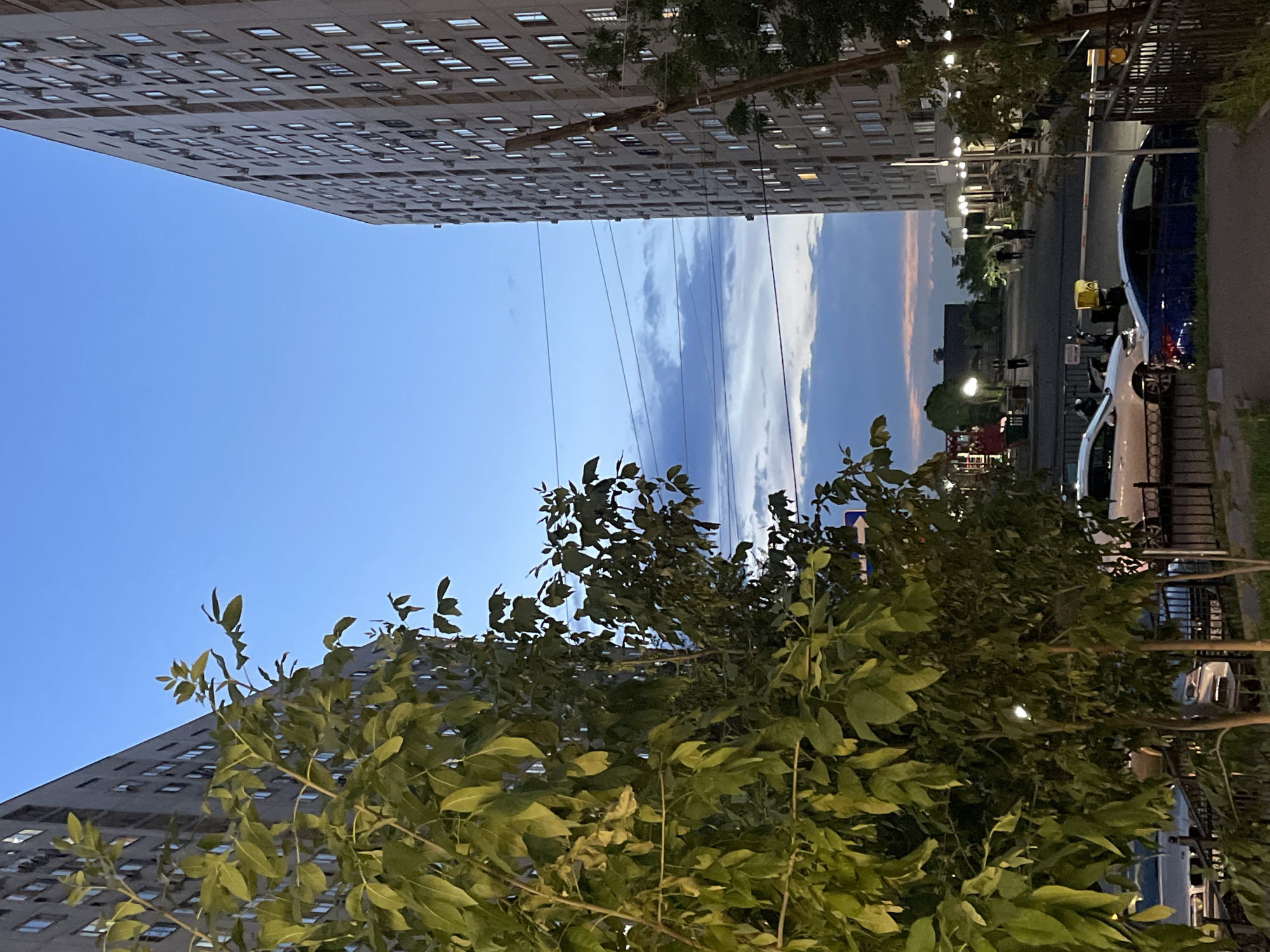 The image depicts an outdoor scene at dusk in Almaty, Kazakhstan, likely in a residential area with high-rise buildings. Two tall, multi-story apartment buildings flank a central courtyard or street. The building on the left is closer to the foreground and partially obscured by lush green trees with dense foliage. The building on the right is further away and shows many lit windows, indicating occupancy. The sky is a gradient of deep blue at the top, transitioning to lighter blue and tinged with pink and orange hues near the horizon, suggesting the afterglow of sunset. Several power lines are visible stretching horizontally across the sky between the buildings.
In the courtyard area, several vehicles are parked, including a white sedan and a dark blue sedan in the foreground, along with motorcycles. A black metal fence runs along the edge of the property. In the middle ground, people are visible walking, some appearing to be conversing or moving about their evening. Streetlights are illuminated, casting pools of light on the ground. Further in the background, there are more buildings and signs of urban infrastructure. The overall atmosphere is one of a typical evening in a city environment.