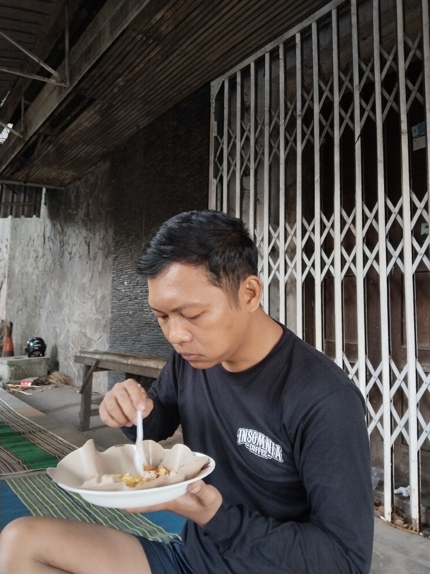 A man in a black long-sleeved shirt with the text "INSOMNIA COFFEE" is seated outdoors, eating from a paper plate with a plastic fork. He is positioned in the foreground of the image. Behind him, a textured stone wall is visible to his left, with a wooden table and a black helmet on a concrete block. To his right, a white accordion-style metal gate covers a wooden door. The ground is covered with striped mats in green, blue, and white. The ceiling above is constructed with dark wooden planks. The scene appears to be an outdoor eatery in Kecamatan Pati, Indonesia.