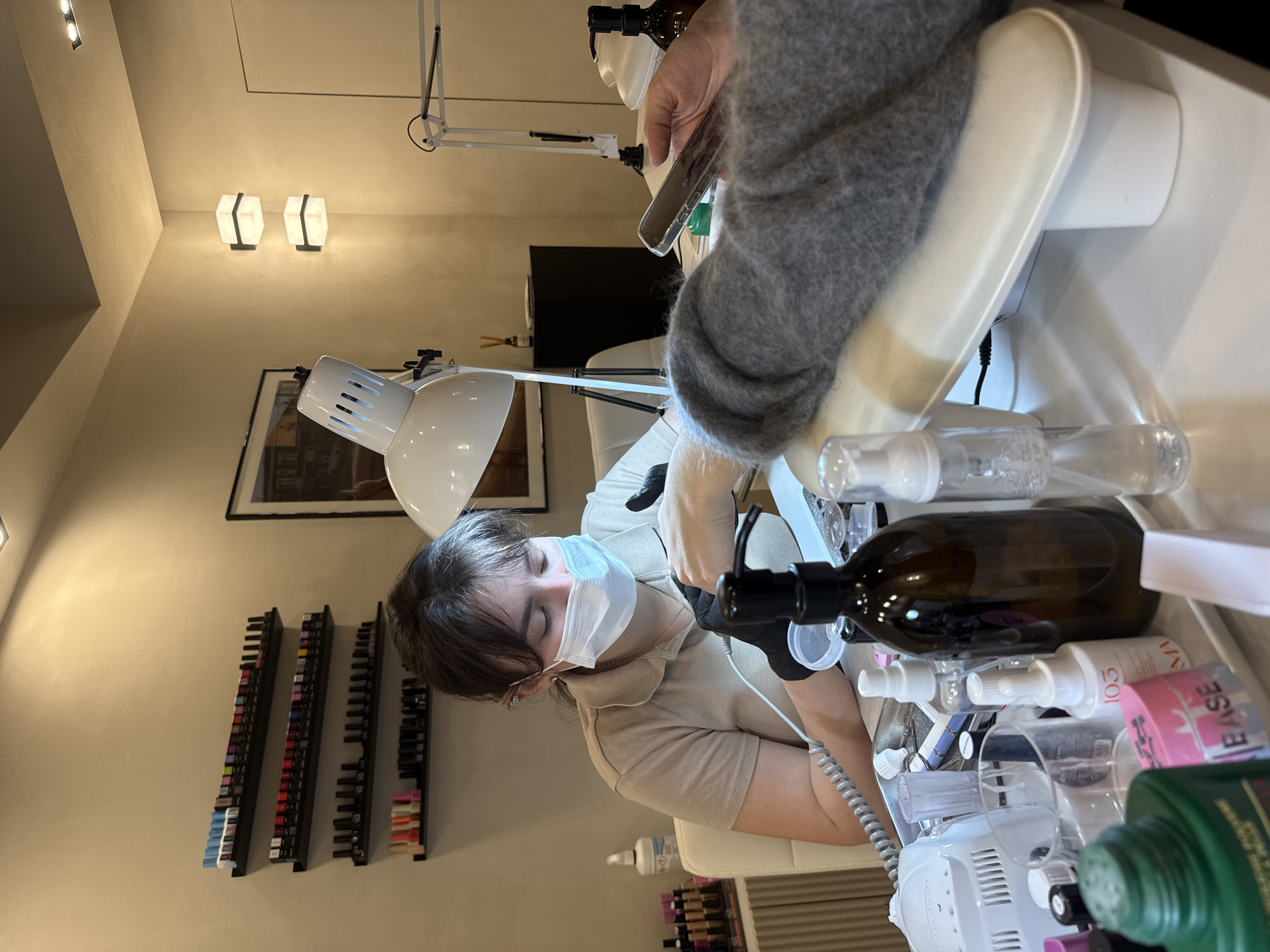 This image, rotated 90 degrees counter-clockwise, captures an indoor scene within what appears to be a nail salon or beauty studio. The primary subjects are a nail technician and a client.
The technician, positioned at the center, is a young woman with brown hair, wearing a light-colored, short-sleeved top, black gloves, and a light blue medical mask. She is leaning over a work area, actively engaged in performing a nail service. Her focus is directed downwards towards the client's hand.
The client's arm and hand are visible on the right side of the frame, clad in a gray knitted sweater. The client holds a smartphone, suggesting they are occupied while receiving the service.
The setting is characterized by light-colored walls, possibly cream or beige. On the left wall, three long, narrow shelves are mounted, displaying numerous rows of nail polish bottles or color samples. A framed picture hangs on the wall behind the technician. Lighting is provided by a large white articulated desk lamp illuminating the workspace, and two modern, rectangular wall-mounted lights with frosted diffusers are visible higher on the background wall.
The white table where the service is being performed is laden with various nail care products and tools. Notable objects include a large dark brown glass bottle with a black pump dispenser, several clear and white spray bottles, and a white electronic device, possibly a nail drill or UV/LED lamp, with wires extending from it. A green bottle is partially visible on the far right. Text "BASE" is clearly visible on a pink container, and "103" and "MA" are readable on a white spray bottle. The overall impression is a professional, well-equipped environment for nail care services. The time of day is not discernible due to the indoor artificial lighting.