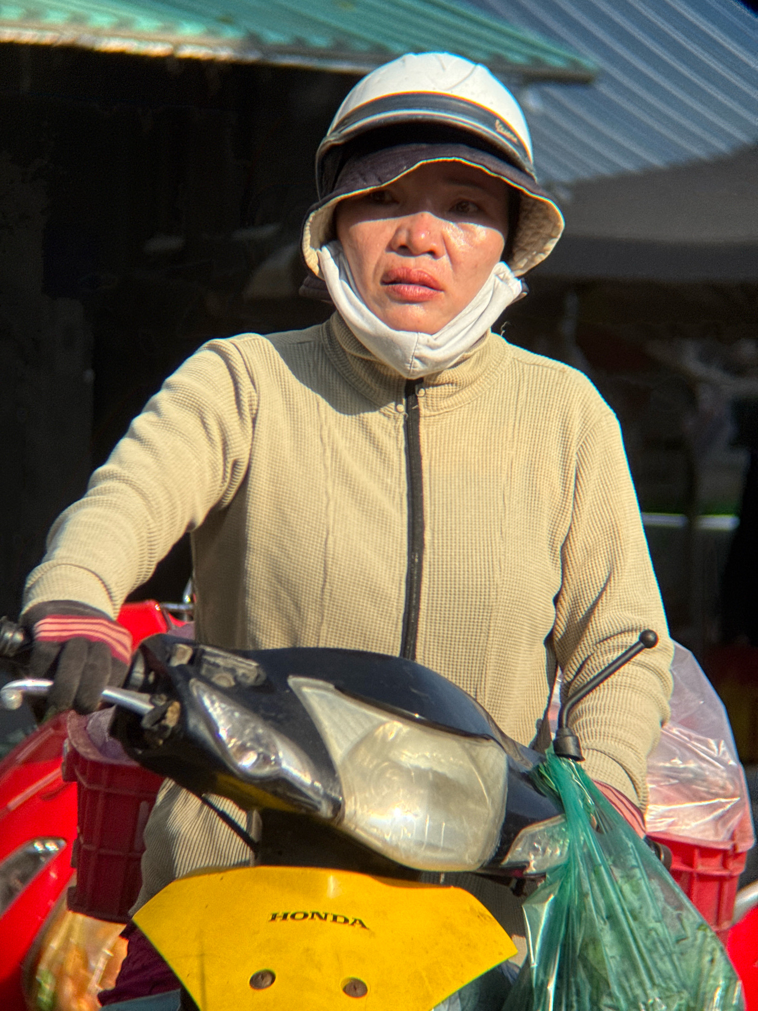 A Vietnamese woman is shown from the chest up, wearing a beige waffle-knit jacket, a white face mask, and a white and dark brimmed hat. She is seated on a scooter, her hands gloved in black with red stripes. The scooter's headlight and yellow front panel are visible, with the "HONDA" logo clearly displayed. A green plastic bag hangs from the handlebars. The background appears to be an outdoor market or street scene with a corrugated metal roof and dark shadows, suggesting a commercial area within Nha Trang, Vietnam.