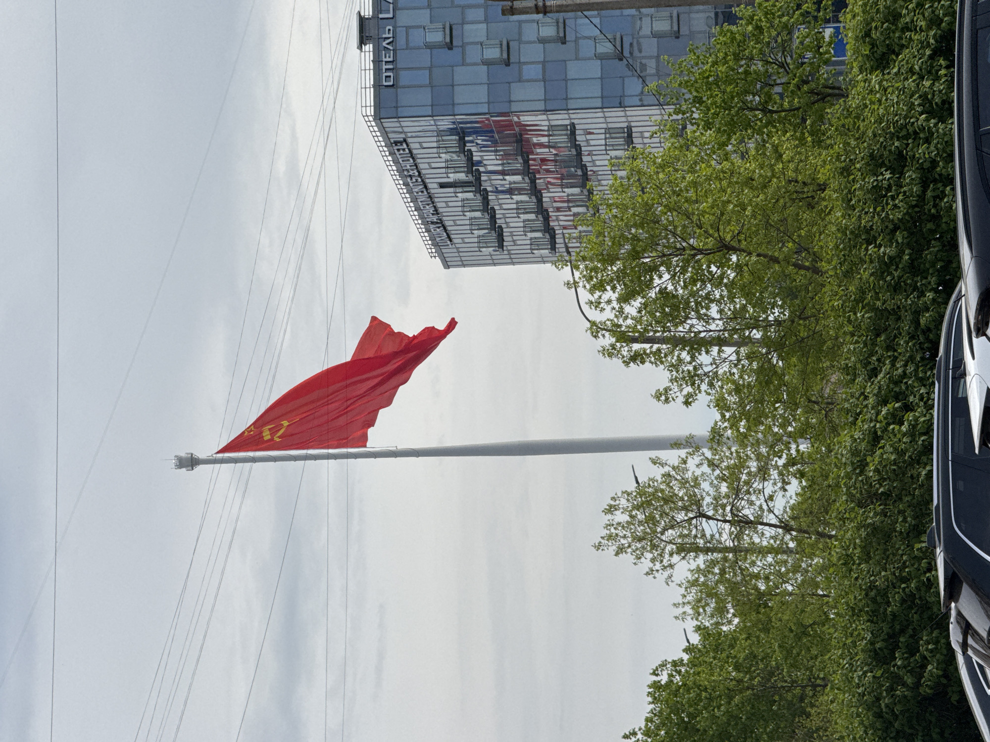 A slightly rotated eye-level shot shows a red flag with a gold design, possibly the flag of the Soviet Union, waving on a long, grey pole. The sky is overcast. In the background, a modern glass building with the Russian word "ОТЕЛЬ" (Hotel) on top is visible. Green leafy trees are to the right of the pole. The side of a black car is partially visible on the right edge of the frame. The scene appears to be in Saint Petersburg, Russia, based on the language on the building and general environment.