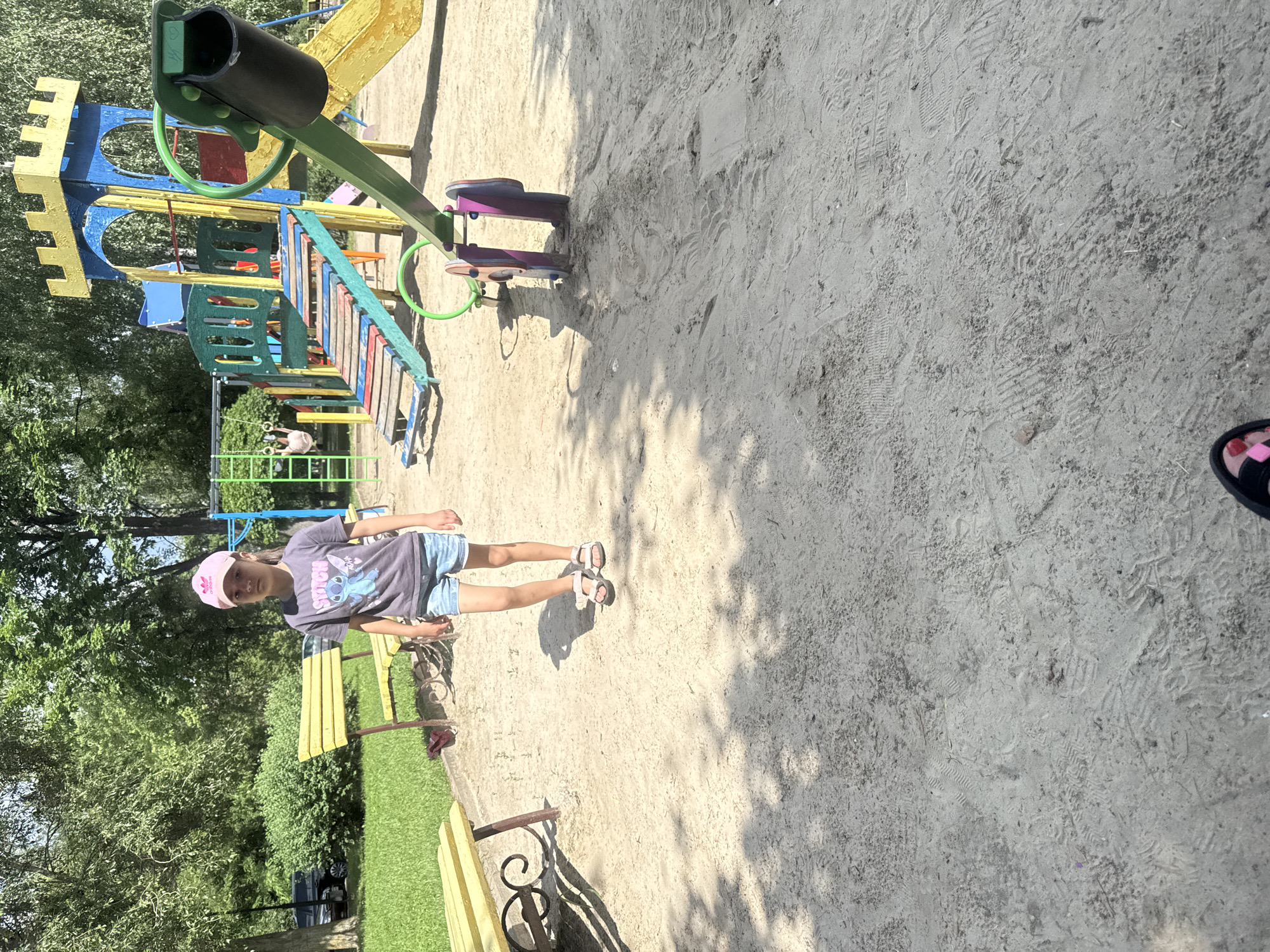 A young girl wearing a pink baseball cap, a dark t-shirt with a Stitch graphic, and denim shorts stands on a sandy playground in Poltava, Ukraine. She is facing forward, looking at the camera with a neutral expression. Behind her, a colorful playground structure with slides and swings is visible, along with yellow benches and trees. The ground is covered in sand, with visible tire tracks and foot imprints, suggesting recent activity. Sunlight casts shadows across the scene, indicating a bright day.