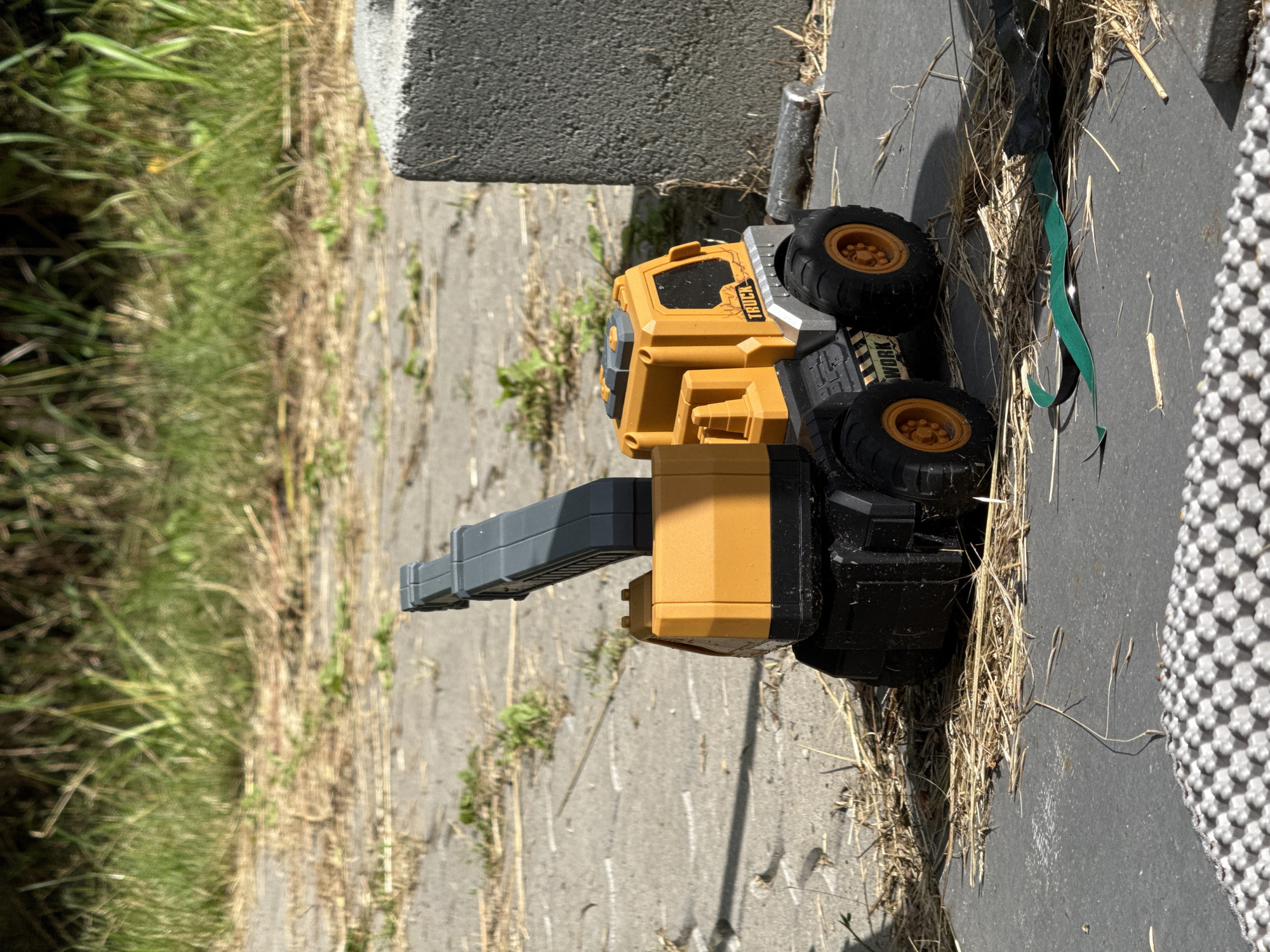 The image shows a yellow and black toy construction truck, likely a digger or excavator, lying on its side on a concrete surface outdoors. The truck is partially covered in dry grass and straw, suggesting it is in a yard or undeveloped area. To the left of the truck is a patch of green grass, and behind it is a grey concrete block. The truck has four large black tires with orange rims. The body of the truck is primarily yellow, with some black and grey components, including a grey arm or boom. On the side of the truck's cabin, the word "TRUCK" is visible in black text. The scene appears to be outdoors, with natural light indicating it is daytime. The presence of dry grass and straw might suggest a warm or dry season. There is a dark green ribbon or strip of material near the truck, partially obscured by grass. The overall setting appears to be a relatively unkempt outdoor area, possibly a backyard or a construction site.