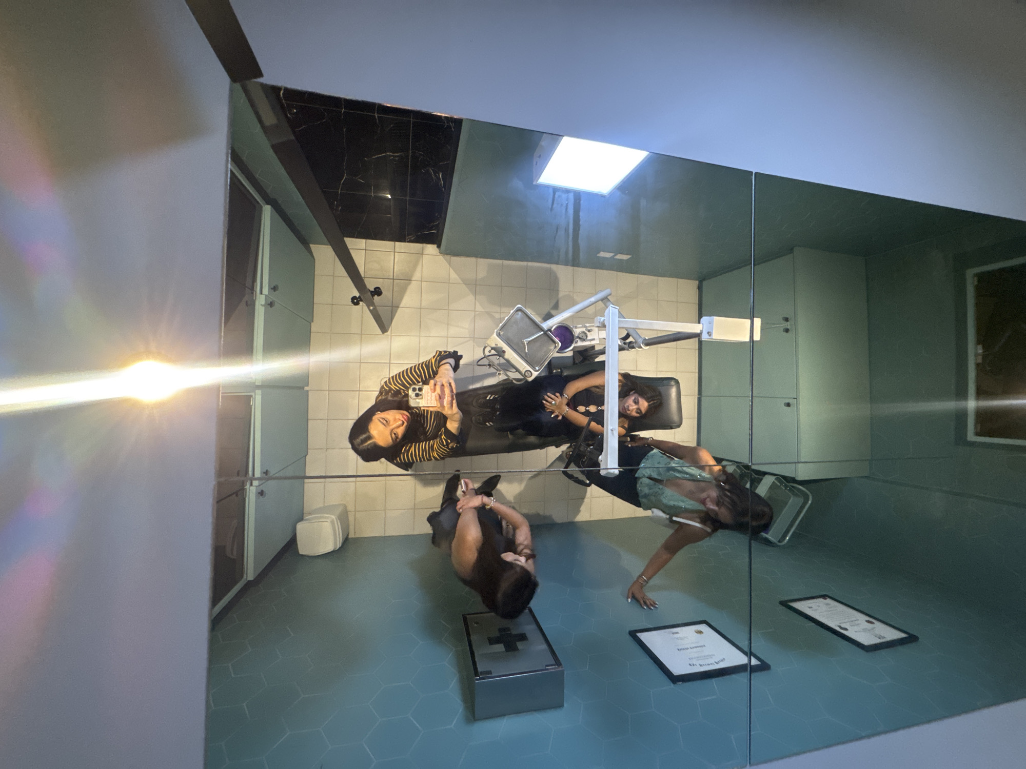 The image depicts an interior room reflected in a mirror, featuring three women in different positions and activities.
One woman is lying on a dental or medical examination chair, seemingly undergoing a procedure or examination. A second woman stands beside the chair, holding a phone and possibly taking a photo of the scene. A third woman is sitting on a silver medical supply or storage box, with two framed certificates visible on the floor nearby.
The room has light blue hexagonal floor tiles, white tiled walls, and some areas of black marble or stone. The lighting appears to be a combination of natural light from a skylight and artificial light. The general appearance suggests a medical or dental office or clinic, possibly modern or recently renovated.
It is not possible to confirm the location as San Bernardino Tlaxcalancingo, Mexico, based solely on the visual information provided in the image.