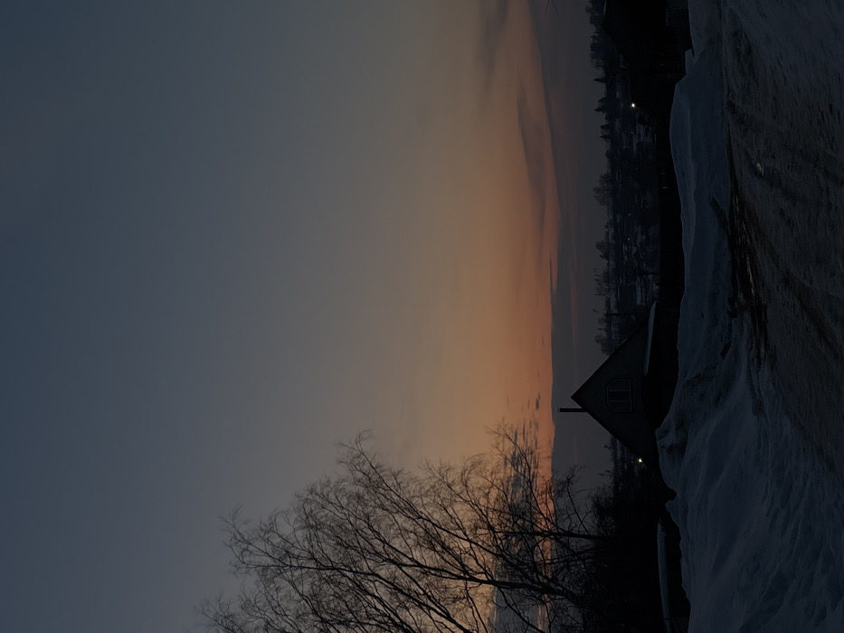 The image captures a winter twilight scene in the city of Фоки, Russia. The upper sky displays a dark grey-blue hue, transitioning to a gradient of orange and reddish tones near the horizon, with several dark, elongated cloud formations present. In the foreground, a snow-covered landscape reveals a dirt track with visible tire impressions and darker, exposed patches. Banks of snow flank both sides of this track. A large, bare deciduous tree, consistent with a birch, is prominently silhouetted on the left, its branches extending into the upper-left quadrant. In the midground, a dark A-frame house, featuring a chimney and an arched window on its gable end, stands silhouetted against the lighter sky. The background shows a distant cluster of buildings and sparse trees forming a settlement, with several small, bright artificial lights visible. No individuals or specific interactions are discernible within the frame.