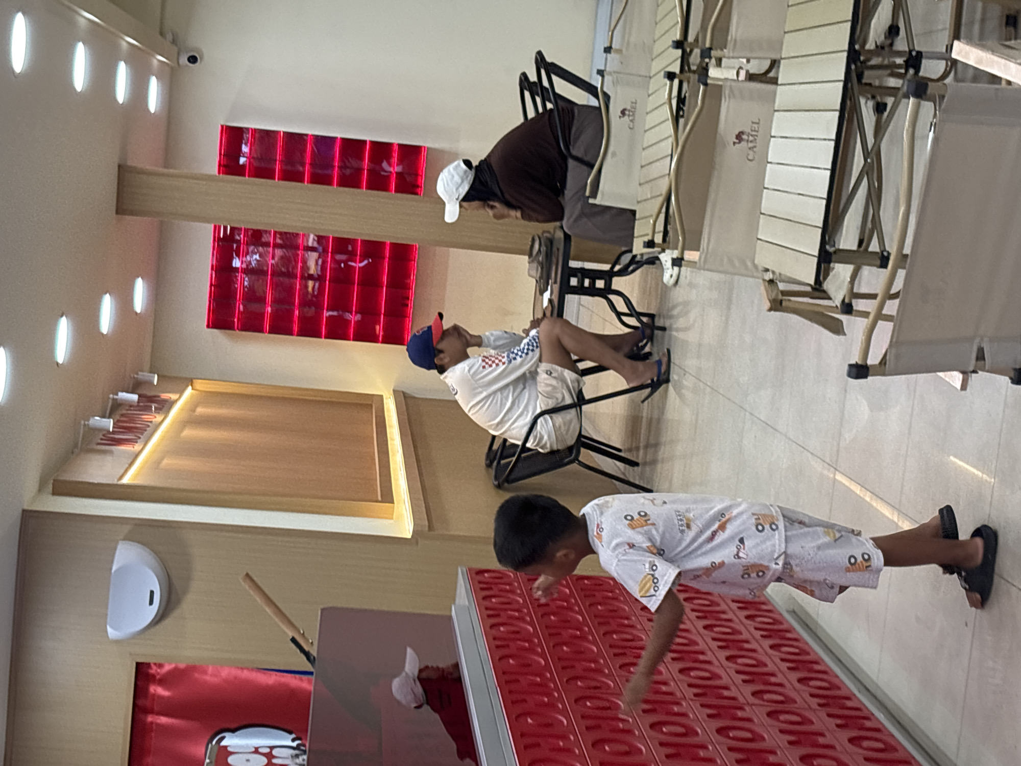 In a brightly lit indoor space, possibly a retail store, located in North Jakarta, Indonesia, three individuals are present. A young boy in a white shirt with cartoon designs and checkered shorts interacts with a red, horizontally oriented object displaying repetitive text. Meanwhile, an adult wearing a brown shirt and a white cap sits in a chair, seemingly focused on something in the foreground. Another person, dressed in a white shirt and checkered shorts with a blue cap, sits in a chair near the adult. The environment includes camping chairs and a wall featuring red-paneled designs and a display case, creating a commercial ambiance.