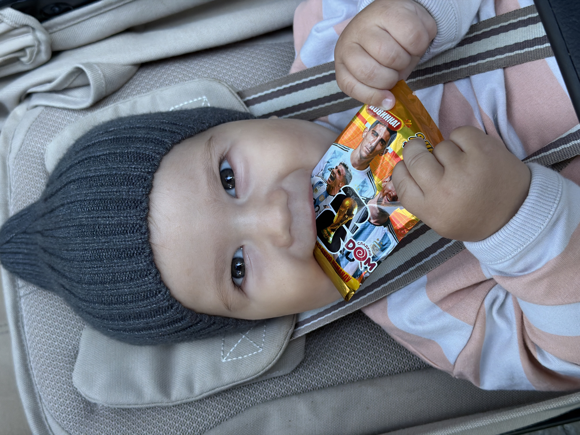 A close-up, overhead shot shows a baby in a stroller, wearing a grey knit hat and a pink and white striped long-sleeved shirt. The baby is holding a small, gold-colored candy wrapper in both hands and is biting on it. The wrapper has colorful graphics and text, including images of soccer players. The baby's face is partially obscured by the wrapper. The background consists of the stroller's fabric in neutral tones. The image does not contain any information about the location of the city of Kazan, Russia.