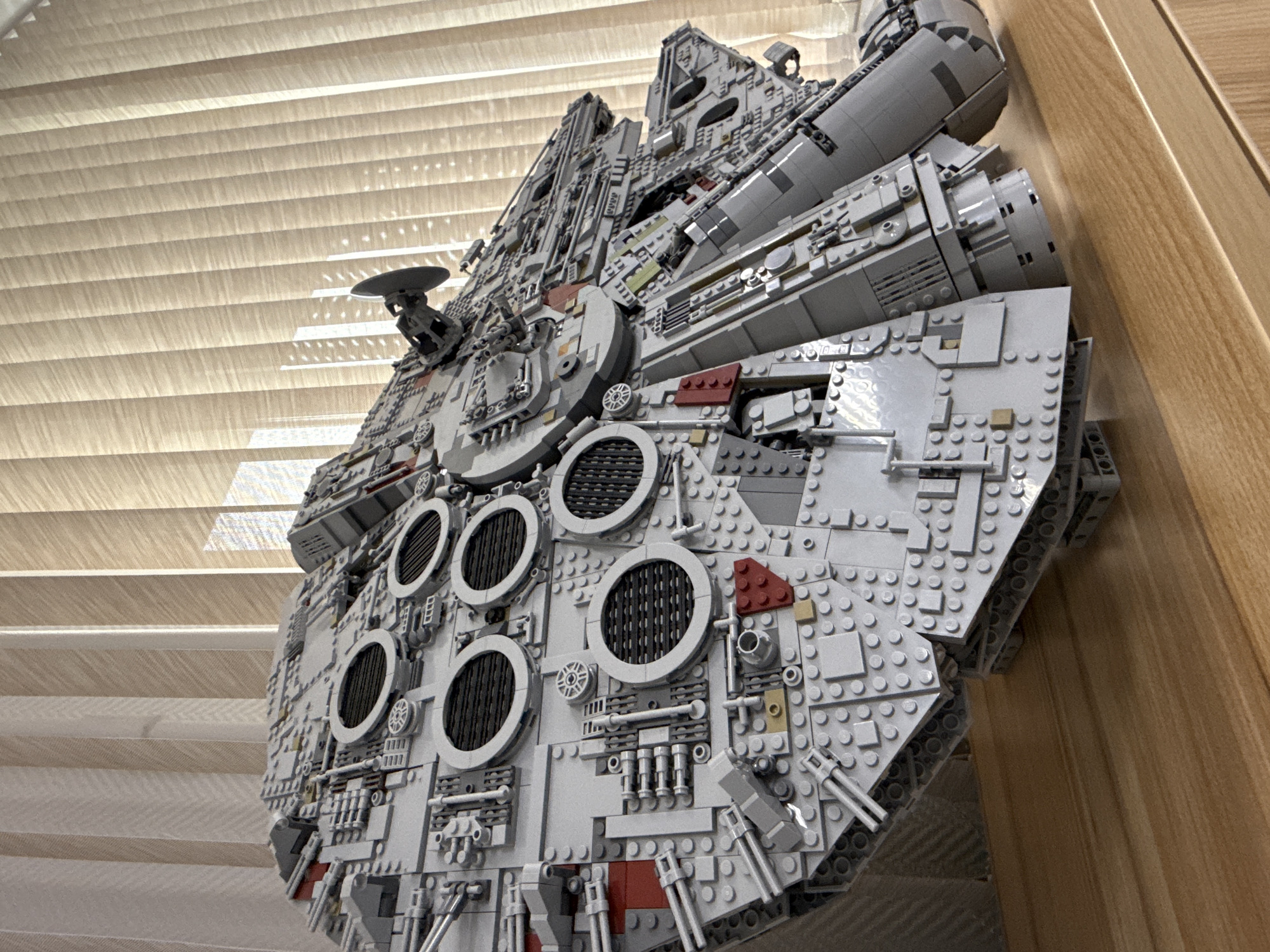 A low-angle close-up shot depicts a Lego Millennium Falcon model. The model is predominantly composed of gray Lego bricks, with details in red and black. The backdrop consists of vertical blinds to the left and wooden paneling to the right. There are no people visible in the image. The model is positioned close to the wall, taking up the majority of the frame. The scene does not provide any information about Krasnoyarsk, Russia.
