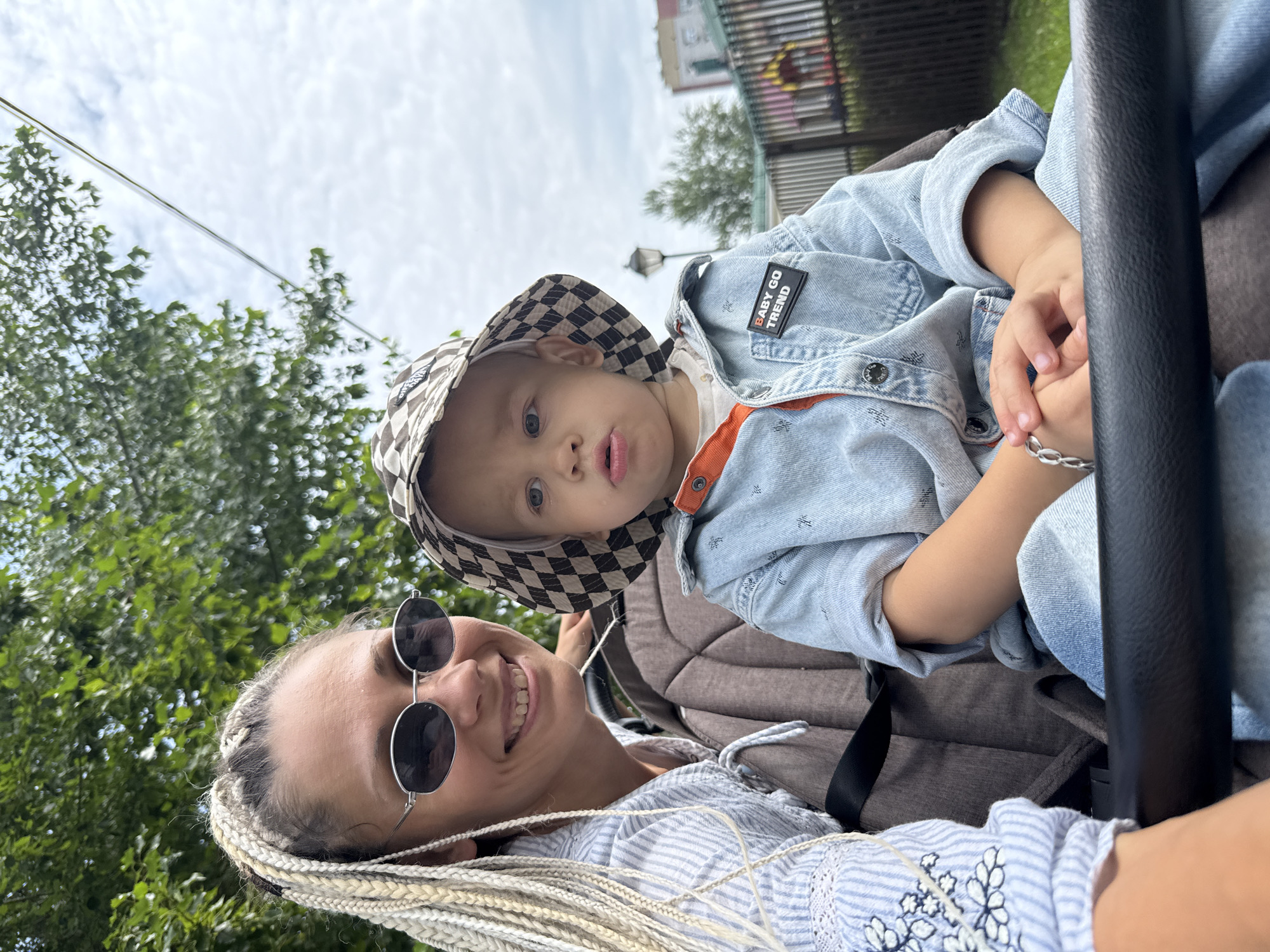 In a medium close-up outdoor shot, a woman with blonde braided hair and sunglasses smiles at the camera while sitting in a stroller. Next to her, a young child wearing a checkered hat and a denim jacket also looks toward the camera. The background shows greenery, a cloudy sky, and buildings with a fence. The image appears to be taken in Rusanovo, Russia.