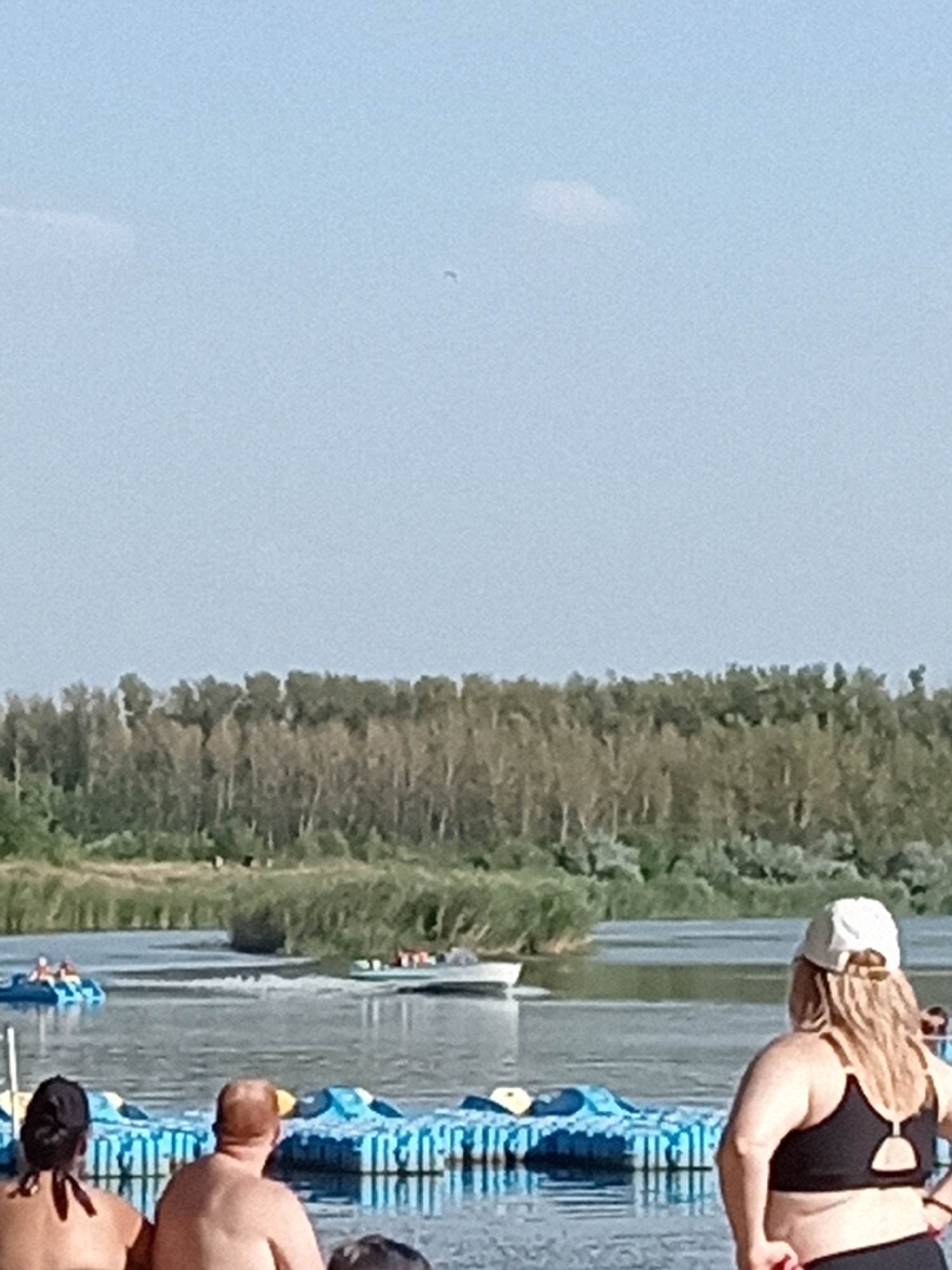 The image shows a sunny day at a lake, likely a recreational area. In the foreground, partially visible, are the backs of three individuals. Two are seated facing away from the camera, one with red hair and the other with dark hair tied up. A third person, wearing a white cap and a black sports bra, stands facing away, looking out at the water. In the middle ground, several people are on the water in what appear to be small, blue pedal boats. In the background, a forested shoreline lines the lake, and the sky is a pale blue with a few wispy clouds. The overall impression is a relaxed summer scene.