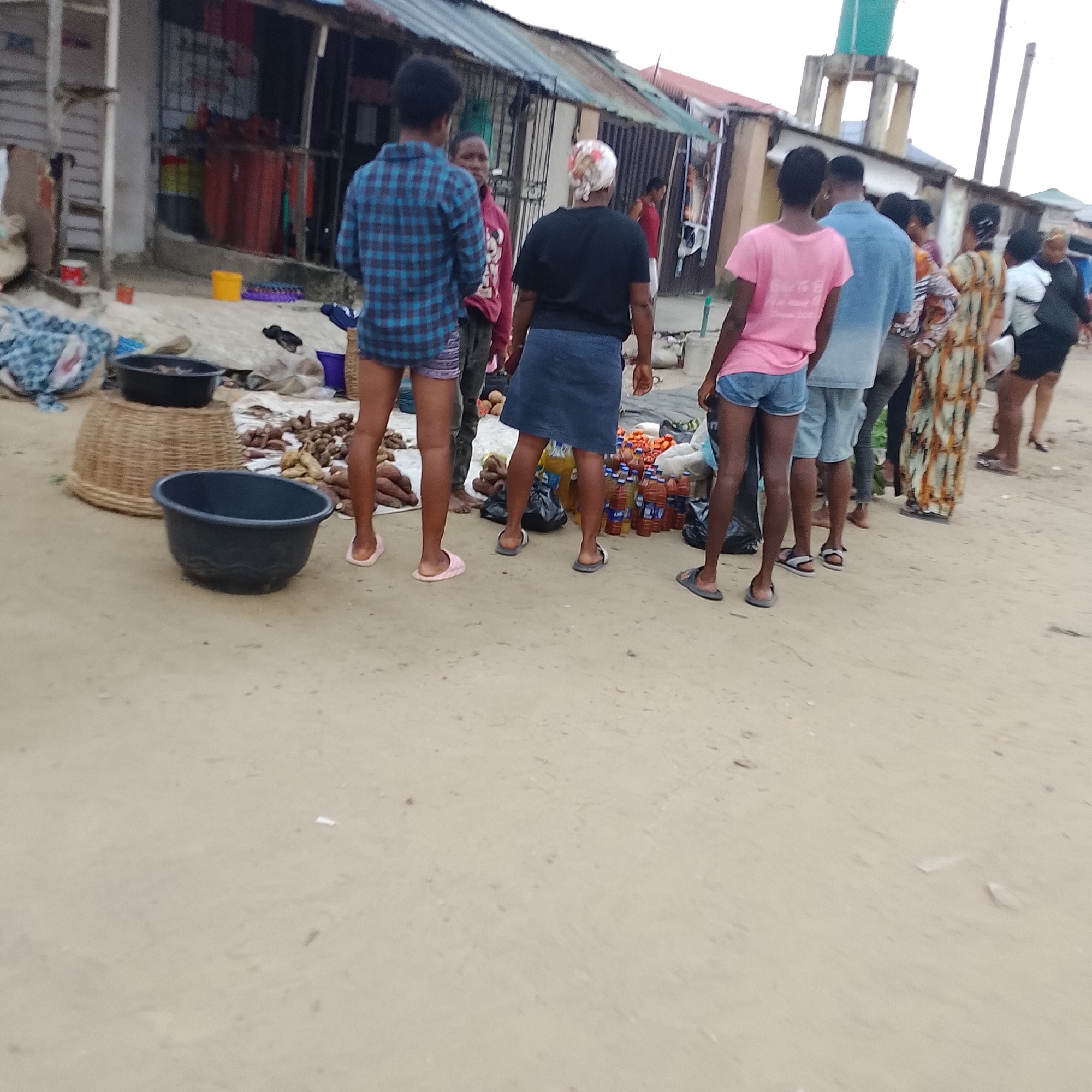 In Ado, Nigeria, a group of people stand around a market stall. The scene unfolds on a dirt ground, with various items for sale and in containers. A woman in a black t-shirt and blue skirt stands near a display of drinks, her back to the camera. To her left, a woman in a pink t-shirt and denim shorts also faces away. A man in a blue button-down shirt and jeans stands in the middle, looking toward the side. Another person in a red hoodie is partially visible behind him. In the foreground, a large black basin sits on the ground, and a woven basket is behind it. A pile of root vegetables, possibly yams or cassava, is spread out on a white cloth. The background features a row of market stalls with corrugated metal roofs and various structures. The overall impression is a bustling, informal marketplace.