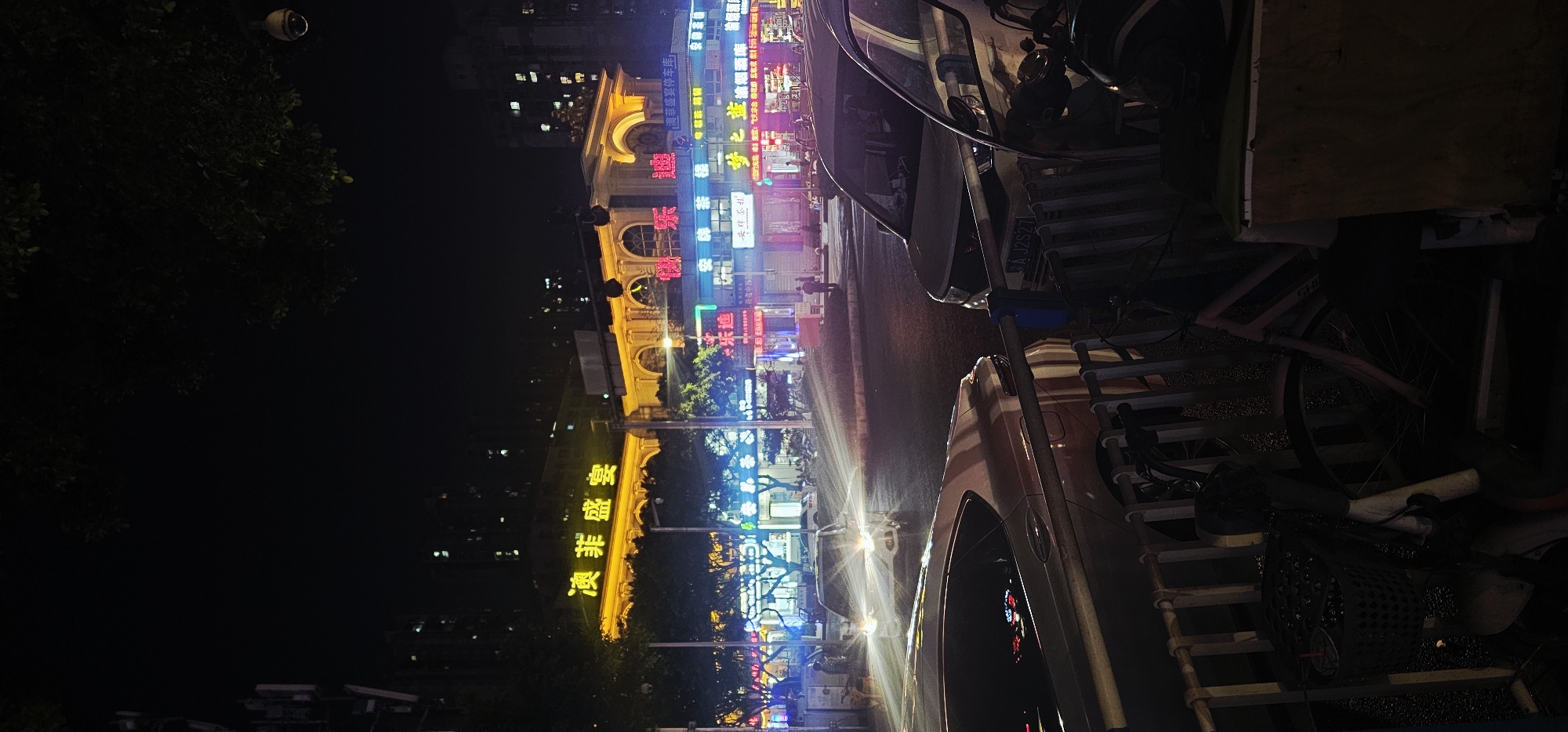 The image shows a nighttime scene in what appears to be the Liangjiang New Area of Chongqing, China. It is a street view with several brightly lit buildings and neon signs written in Chinese characters. Some of the signs read "澳菲盛宴" (Aofeishengyan, possibly a banquet hall), "澳非盛宴停车库" (Aofeishengyan Parking Garage), and "安牌茶糕" (Anpai Chagao). There's also a sign for "梦之蓝烟" (Mengzhilan Yan). Cars are parked along the street, with a car license plate "A J2S27" visible. There is street lighting, and blurred figures of people are present in the background. The foreground contains cluttered items, including what appears to be bicycles and ladders. The lighting is vibrant and typical of an urban night scene in China.