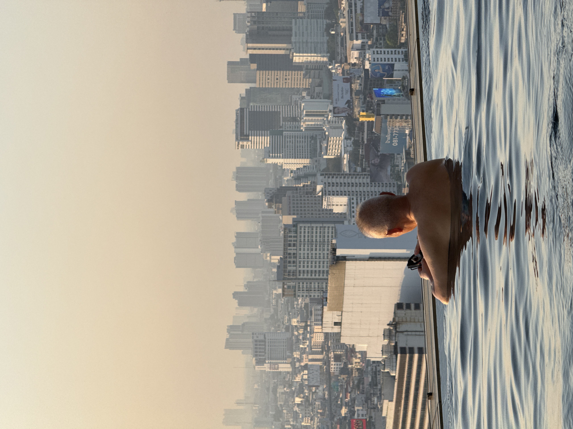 A high-angle, outdoor shot shows a bald, light-skinned person in a swimming pool with a cityscape background. The subject is in the right side of the frame, and facing toward the cityscape. The water's surface is reflecting the light. The cityscape is comprised of many high-rise buildings with a light haze. The location appears to be Bangkok, Thailand.