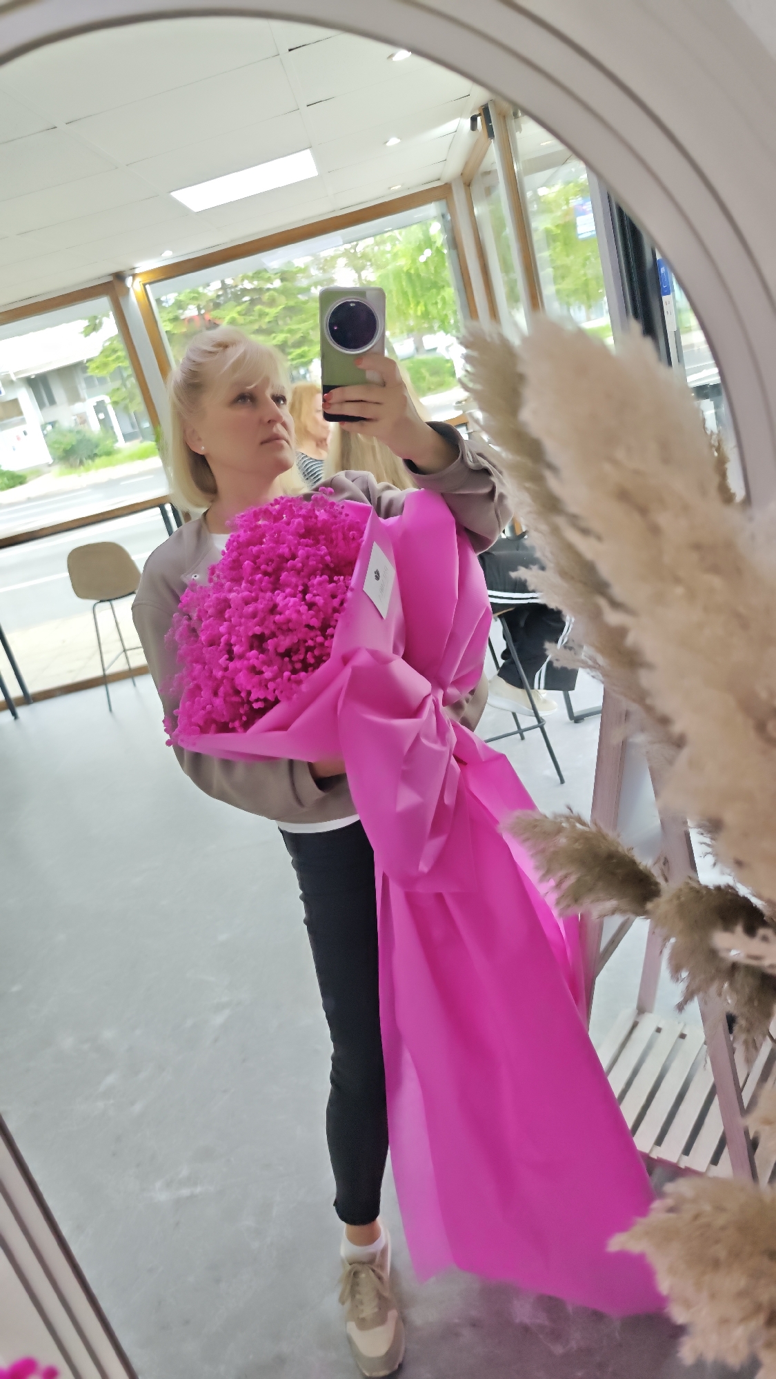 A woman is taking a mirror selfie while holding a large bouquet of bright pink flowers wrapped in matching pink paper. The bouquet obscures much of her body, but she is wearing black leggings, a light-colored sweatshirt, and beige sneakers. Her blonde hair is partially pulled back. She is holding a smartphone up to take the picture. The reflection shows a room with large windows looking out onto a city street. There are chairs and other people visible in the background. Fluffy, beige pampas grass decor is on the right side of the mirror, partially obscuring the reflection. Based on the architectural style and street view, it is very likely this photo was taken in Burgas, Bulgaria.