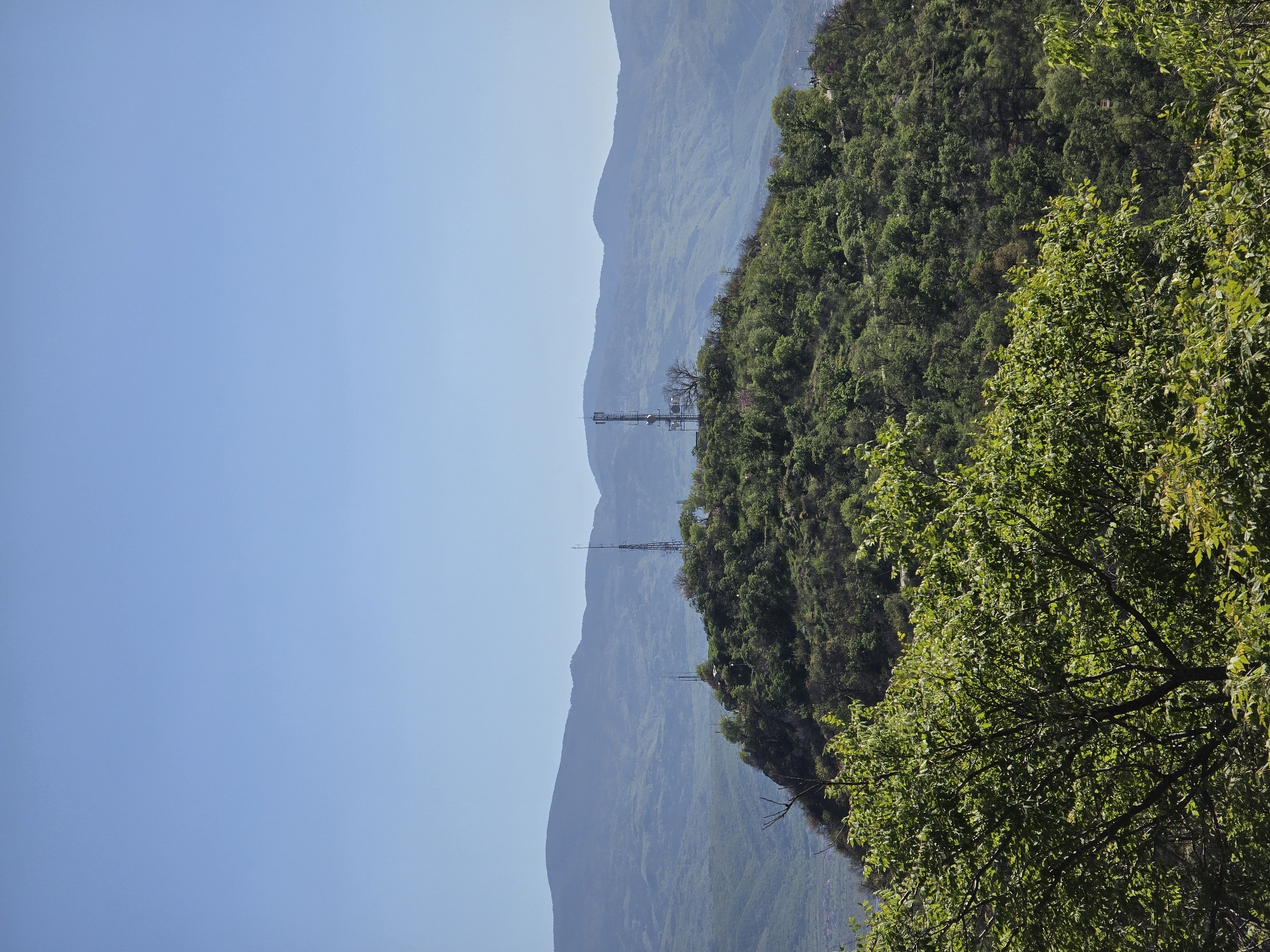 The image depicts a landscape of mountainous terrain with a clear, blue sky on the left side. A series of mountain ranges are visible in the background. Closer to the right, a hillside covered in dense green foliage is present, with a tree in the foreground partially obscuring the view. Communication towers are situated on one of the mountain ranges, indicating the presence of infrastructure. The image's location and specific details about the city are unknown.