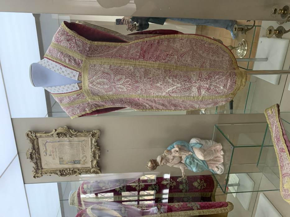The image displays an interior museum exhibit, likely within the Radziwill Castle Museum in Nyasvizh, Belarus. The central object is a sleeveless ecclesiastical vestment, possibly a dalmatic, exhibited on a white mannequin torso. This vestment is predominantly pink or magenta, adorned with intricate gold and white floral and foliate patterned embroidery, and features gold trim along its edges and vertical front panels. The V-neck collar is detailed with a white openwork fabric.
To the upper left, a framed document with an ornate gold Baroque-style frame and handwritten, illuminated script is mounted on a beige wall. Below this framed document, on a clear glass pedestal, rests a polychrome statue depicting the crowned Virgin Mary seated and holding the infant Jesus; Mary wears a blue cloak and a light-colored dress, while Jesus is partially draped.
On the far left, another pink and gold patterned ceremonial garment, possibly a cope, is partially visible within a glass display enclosure, reflecting a window with grid patterns. A strip of matching fabric, possibly a maniple or stole, lies on a lower clear glass shelf in the foreground. In the reflections on the right-hand glass panel, a person holding a smartphone is visible, indicating the act of photography. Partially visible at the bottom right are brass ecclesiastical candle holders. The environment consists of beige walls, glass display cases, and a white ceiling.