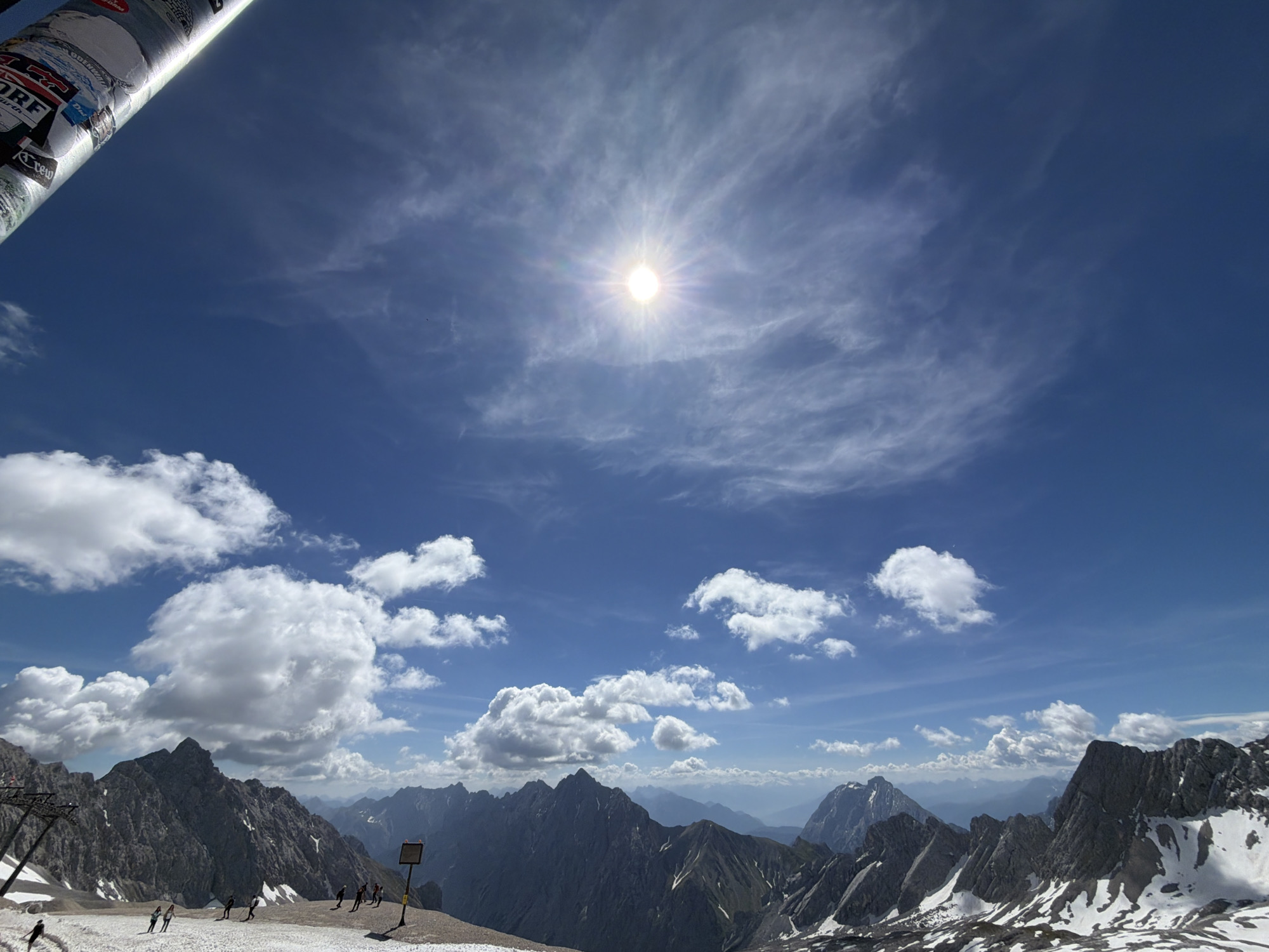 This is an outdoor, wide-angle photograph taken from a high vantage point, likely a mountain peak, overlooking a vast mountain range under a clear blue sky with scattered white clouds. The sun is prominent in the upper-middle of the frame, shining brightly with a lens flare. In the foreground, to the left, a cylindrical object covered in stickers is partially visible, extending from the top left corner downwards. The object appears to be a pole or a railing. Below this, a snowy slope is visible with several people, some appearing to be skiing or snowboarding down the slope. A ski lift structure is also present on the left side. The middle ground and background are dominated by rugged, rocky mountain peaks, some of which are dusted with snow. The distant mountains fade into a hazy blue. The image conveys a sense of a sunny day in a mountainous, possibly ski resort, environment. Given the context of Garmisch-Partenkirchen, Germany, this scene likely depicts the Zugspitze, Germany's highest peak, known for its ski areas and panoramic mountain views.