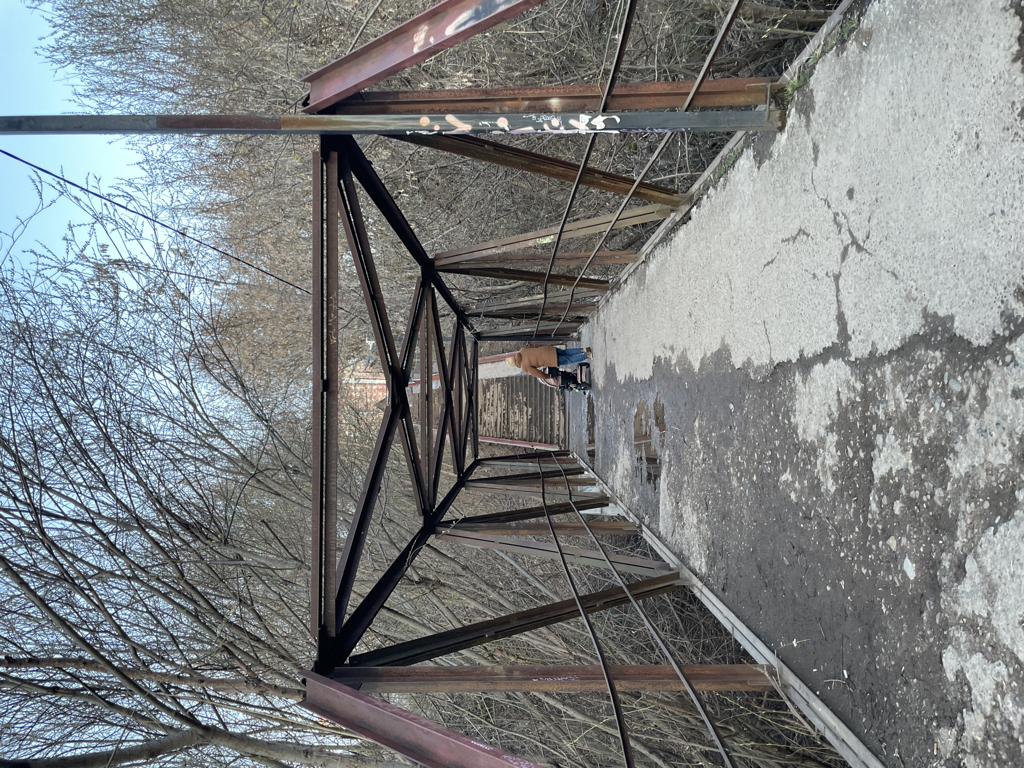 The image depicts a pedestrian bridge viewed lengthwise from one end, looking towards the opposite end. The bridge features a rusty metal truss structure forming an enclosed tunnel-like passage, with a concrete or asphalt walkway.
In the distance, a sole individual is visible walking away from the camera, pushing a stroller or cart. The person is wearing a light brown or tan jacket and blue jeans.
The setting appears to be a natural or semi-natural area, with numerous bare trees on either side of the bridge, their intricate branches visible through and above the metal framework. The sky above is a pale blue, suggesting clear weather during the daytime, likely in late autumn or winter given the deciduous trees are without leaves.
Notable details include the weathered condition of the bridge structure, which shows significant rust and wear. Graffiti is present on several metal beams; on the upper right beam, abstract white and light pink marks are seen, some resembling letters like "OWO". On a lower left beam, a tag that might read "GRUNGE" or "CRINGE" is visible in white, alongside other illegible spray paint. The walkway itself is cracked and discolored in places, with patches of dirt or moss, and some green weeds growing along the edges. Wires or cables run along the interior of the bridge's left side.
The overall impression is one of an aged, utilitarian bridge in a somewhat neglected state, traversing a natural landscape in a cool season. The location context indicates Izhevsk, Russia.